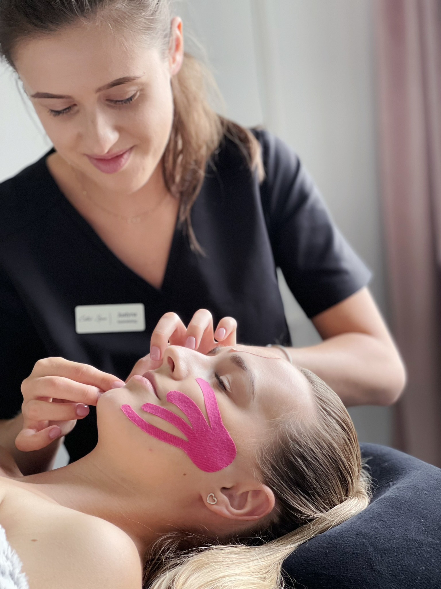 Kobieta poddaje się zabiegowi kinesiotapingu twarzy; różowe paski aplikowane przez kosmetyczkę w czarnym uniformie.