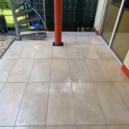 Beżowe płytki ułożone na zewnątrz, tworzące taras z widoczną kolumną pomalowaną na pomarańczowo, obok schody spiralne i żwirowe obrzeże.