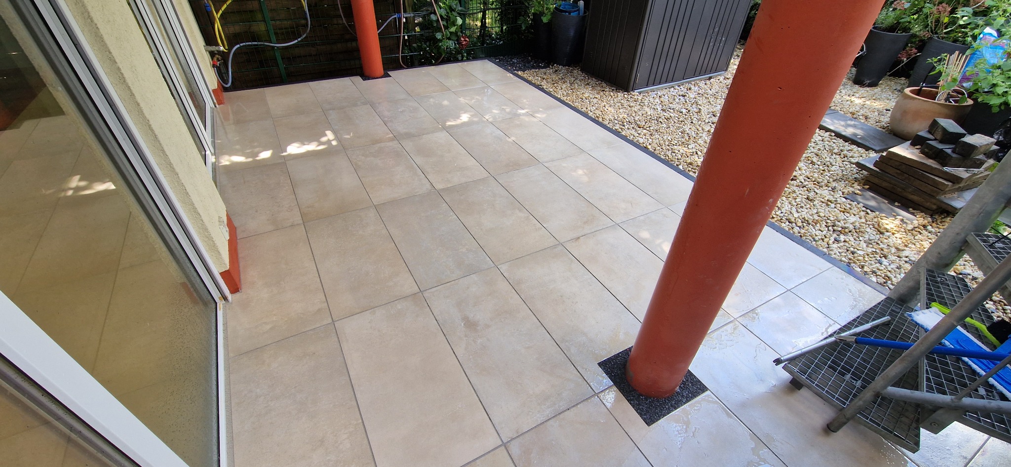 Jasne, kwadratowe płytki podłogowe na zewnątrz, z pomarańczową kolumną i drabiną w rogu, widoczne narzędzia do czyszczenia na drabinie.