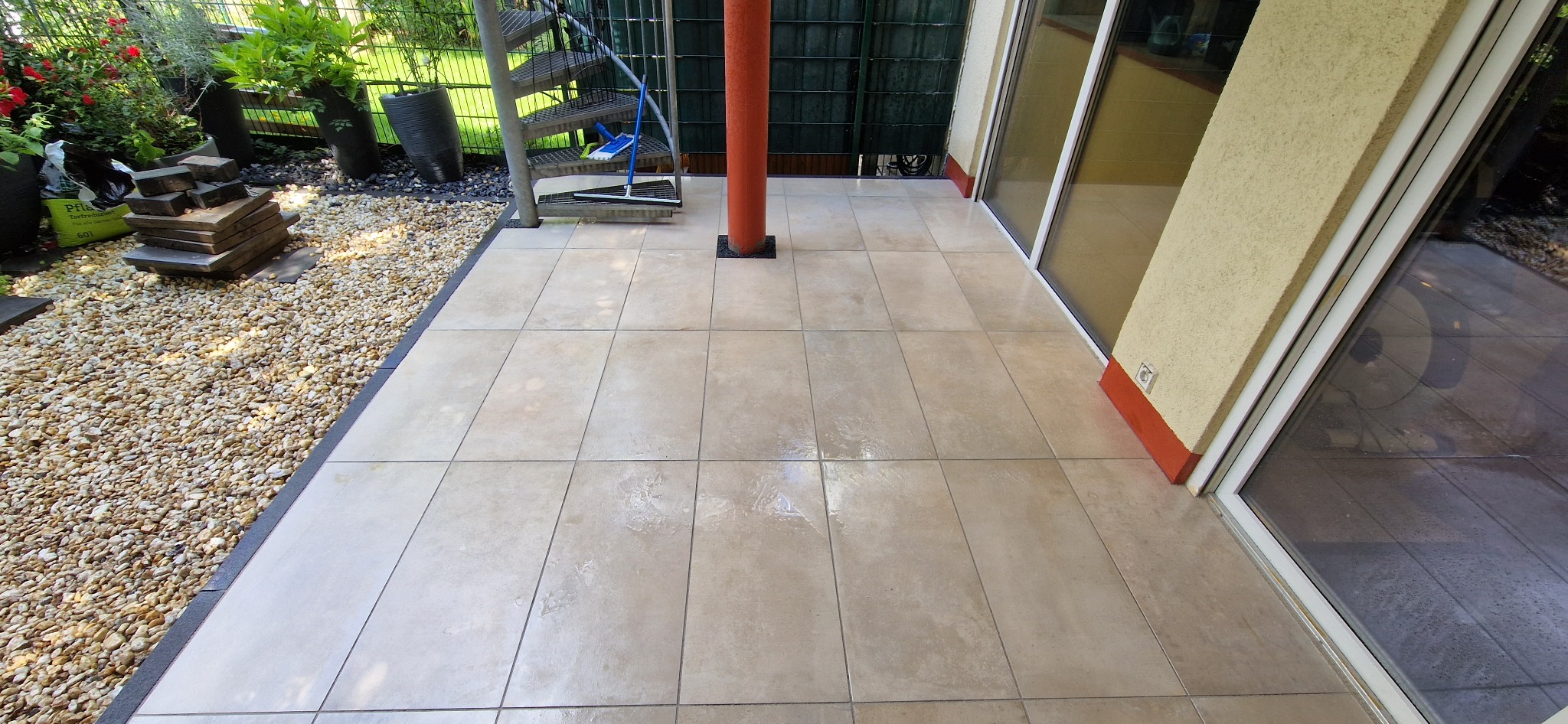 Beżowe płytki ułożone na zewnątrz, tworzące taras z widoczną kolumną pomalowaną na pomarańczowo, obok schody spiralne i żwirowe obrzeże.