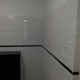 Białe, błyszczące płytki ścienne z delikatnym wzorem fal, oddzielone czarnym pasem, w rogu pomieszczenia.