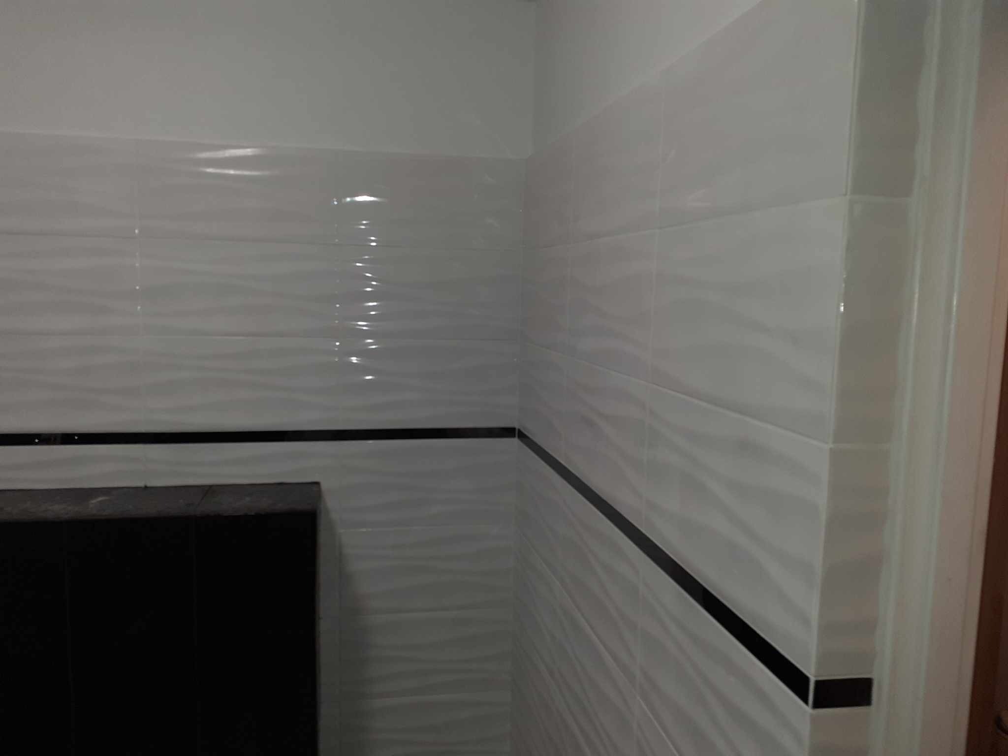 Białe, błyszczące płytki ścienne z delikatnym wzorem fal, oddzielone czarnym pasem, w rogu pomieszczenia.