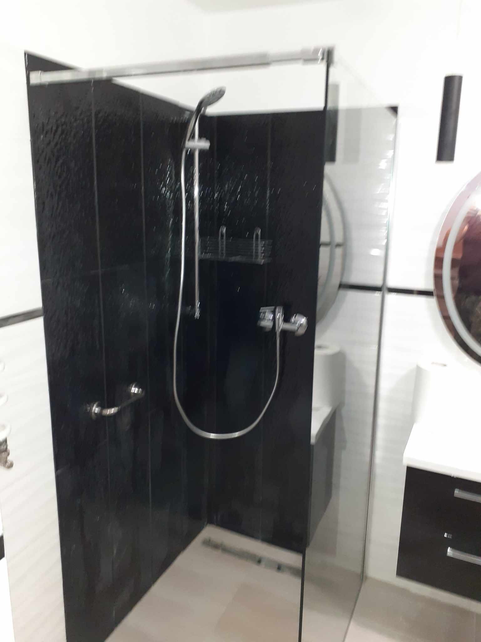 Wykończona kabina prysznicowa z czarnymi płytkami, chromowaną armaturą i szklaną ścianką, widok na umywalkę z szafką w tle.
