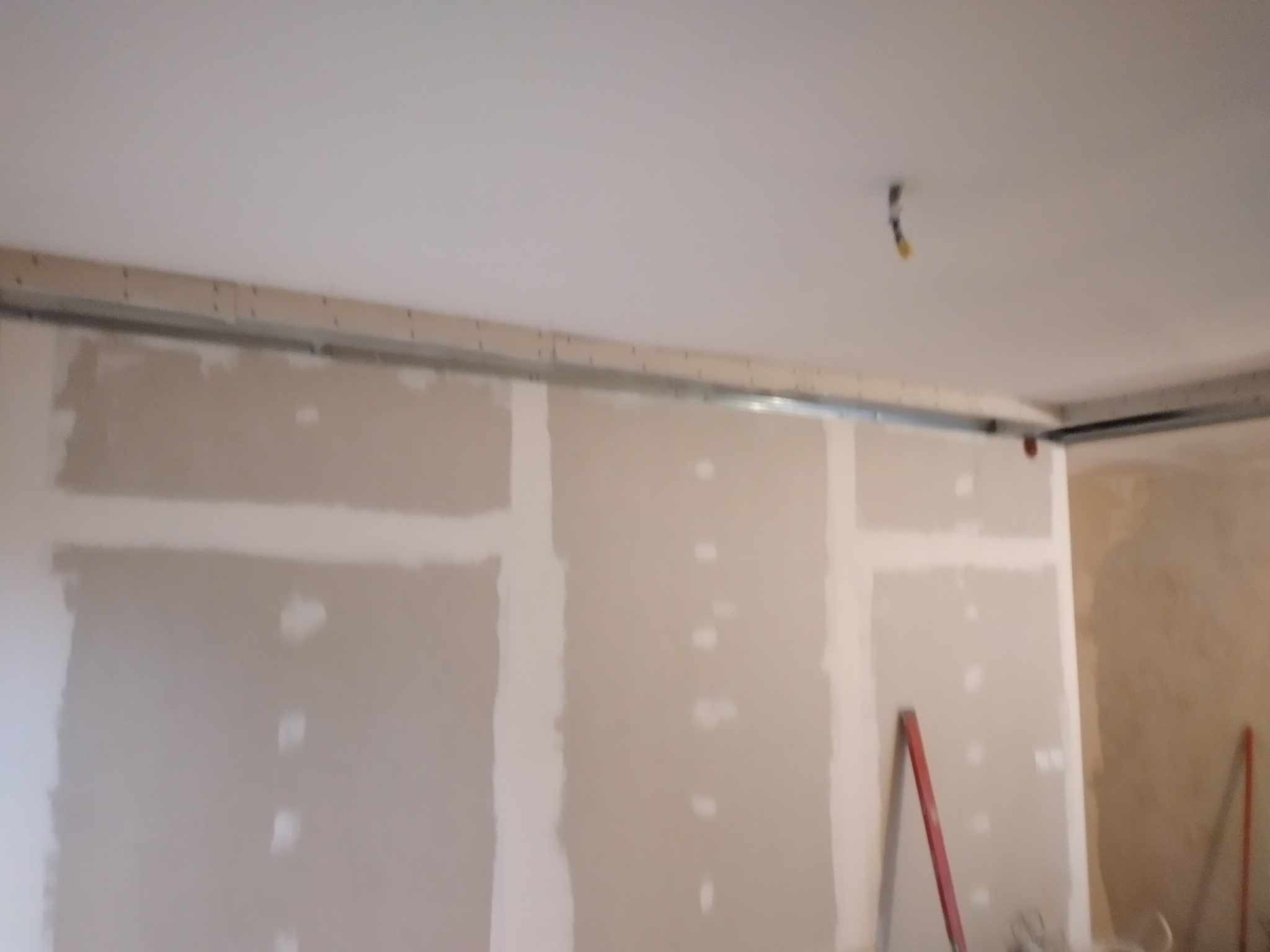 Montaż płyt gipsowo-kartonowych na metalowym stelażu przy ścianie, widoczne szpachlowane łączenia, drabina oparta o ścianę.