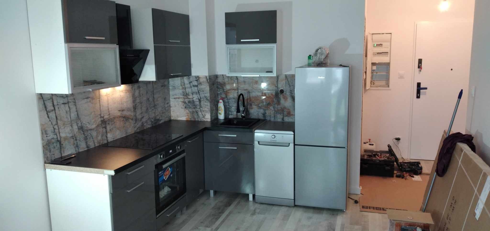 Nowoczesna, szaro-biała kuchnia z marmurowym backsplash w mieszkaniu w Luzinie, widok na zabudowę kuchenną z piekarnikiem, zlewem i lodówką Amica. W tle widoczne narzędzia i kartony.