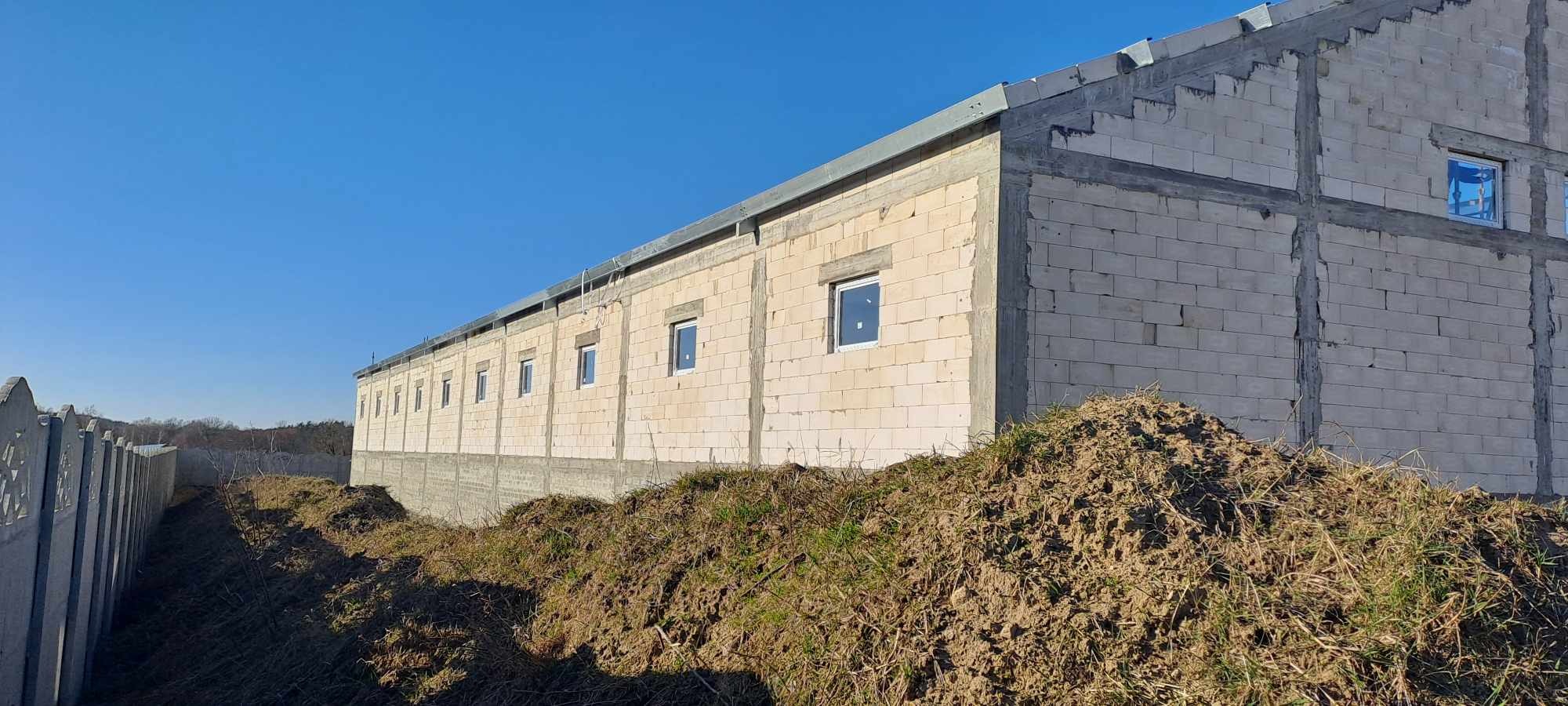 Budynek w trakcie budowy z białych bloczków, widoczne okna, z zewnątrz ziemia i trawa, obok ogrodzenie betonowe.