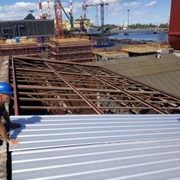 Dom-Dach Domski Mateusz - Pracownicy w kaskach montują blachę trapezową na drewnianej konstrukcji dachu przemysłowego, w tle widoczne dźwigi portowe i nabrzeże.