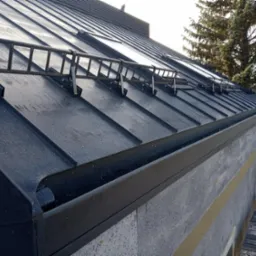 Czarny, metalowy dach z zamontowanymi zabezpieczeniami przeciwśniegowymi w formie drabinki, widoczna również rynna i fragment ocieplonej ściany budynku.