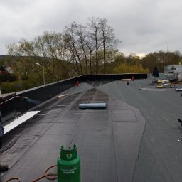 Dom-Dach Domski Mateusz - Prace na płaskim dachu pokrytym czarną papą termozgrzewalną; widoczna butla gazowa Eurogaz, palnik dekarski w użyciu, częściowo położona biała warstwa izolacyjna, oraz narzędzia i materiały...