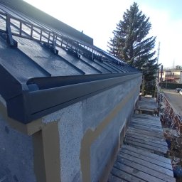 Dom-Dach Domski Mateusz - Montaż czarnej blachy na dachu z zamontowanymi drabinkami przeciwśniegowymi, widoczne rusztowanie i ocieplenie budynku w trakcie prac wykończeniowych.