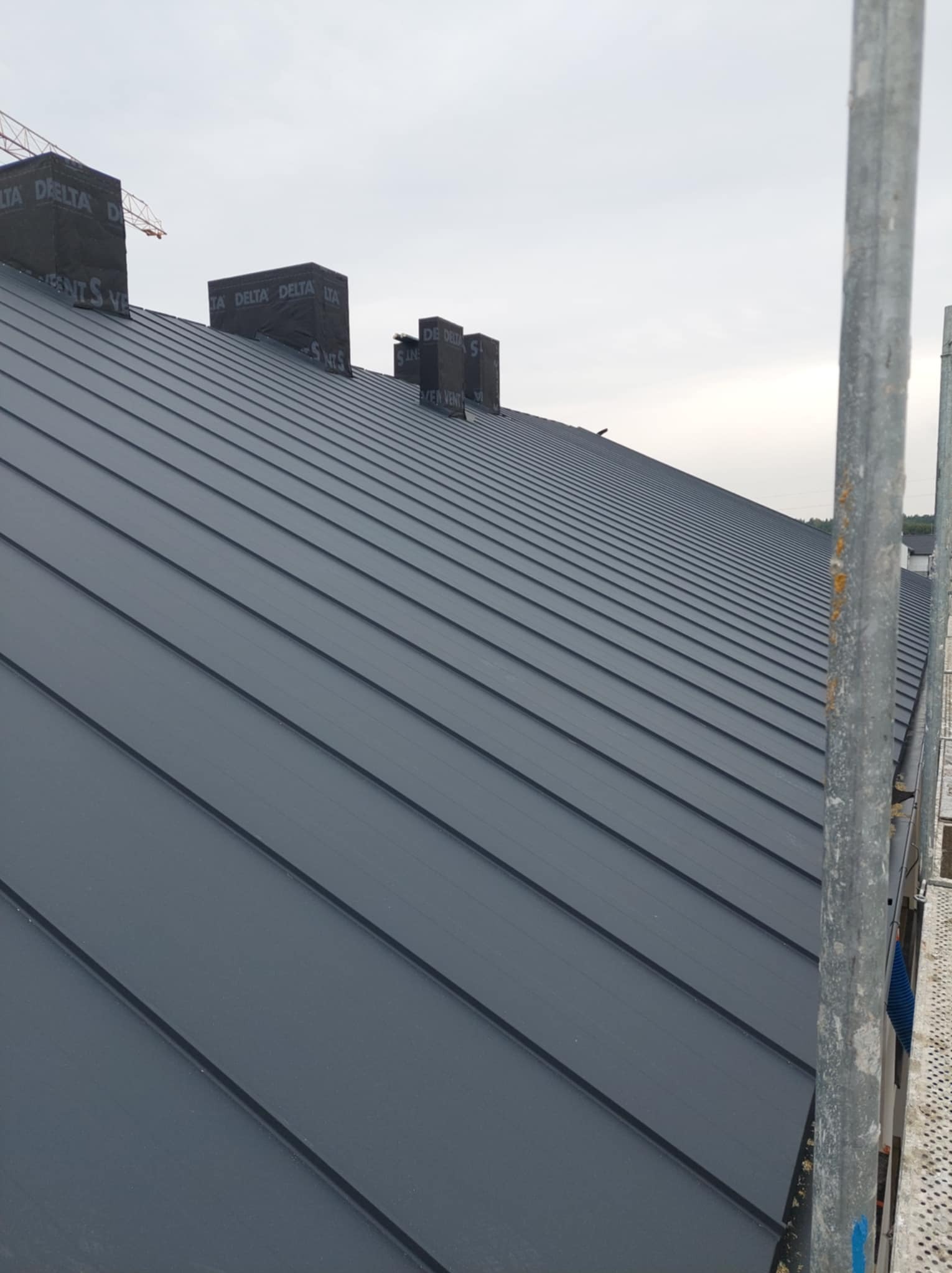 Szary dach z blachy na rąbek stojący, widoczne kominy zabezpieczone czarną folią Delta, w tle żuraw budowlany i pochmurne niebo, ujęcie ze rusztowania.
