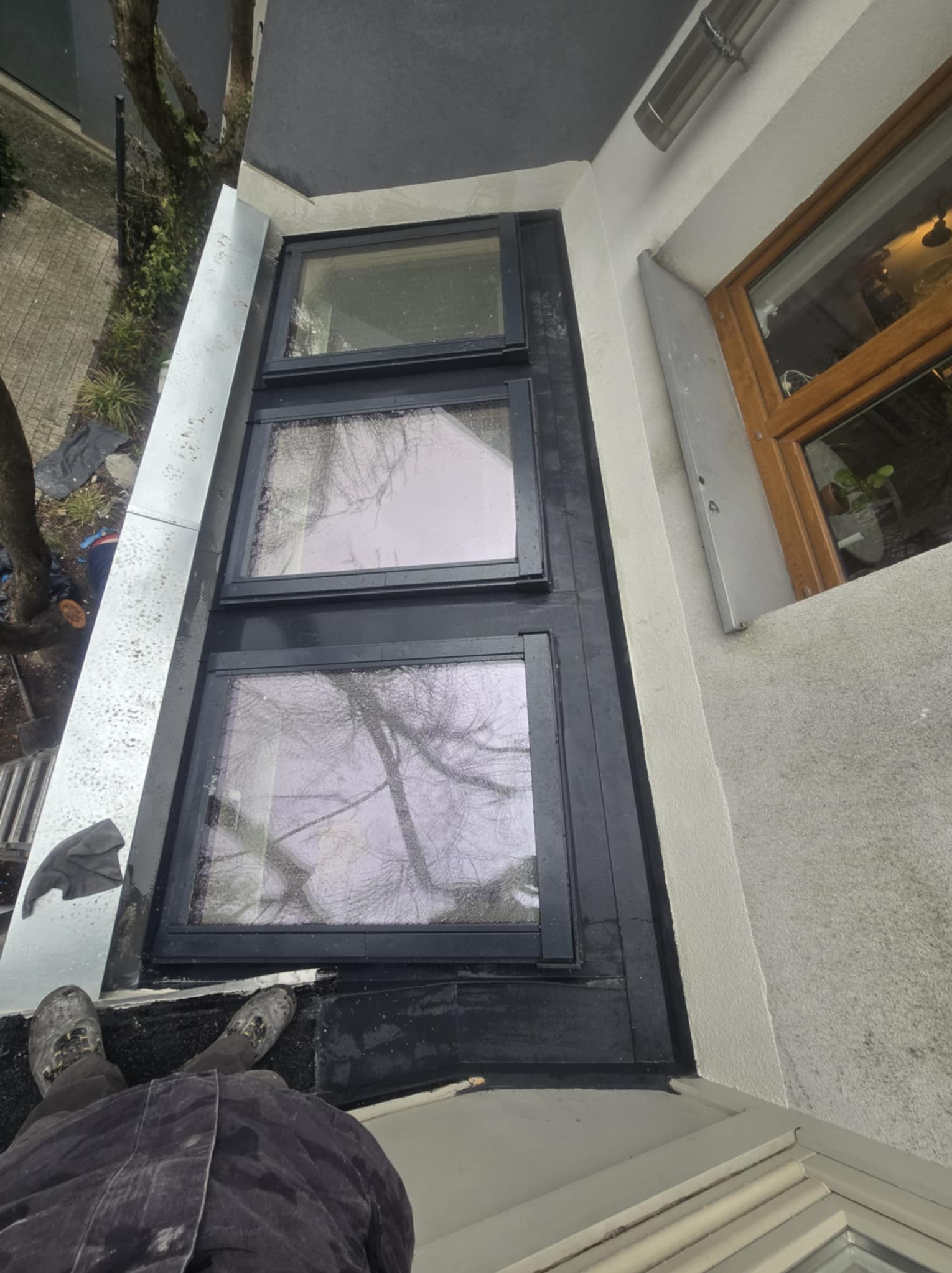 Widok z góry na trzy okna dachowe z czarnymi ramami, zamontowane na płaskim dachu pokrytym czarną papą, z widocznymi obróbkami blacharskimi i fragmentem rynny.