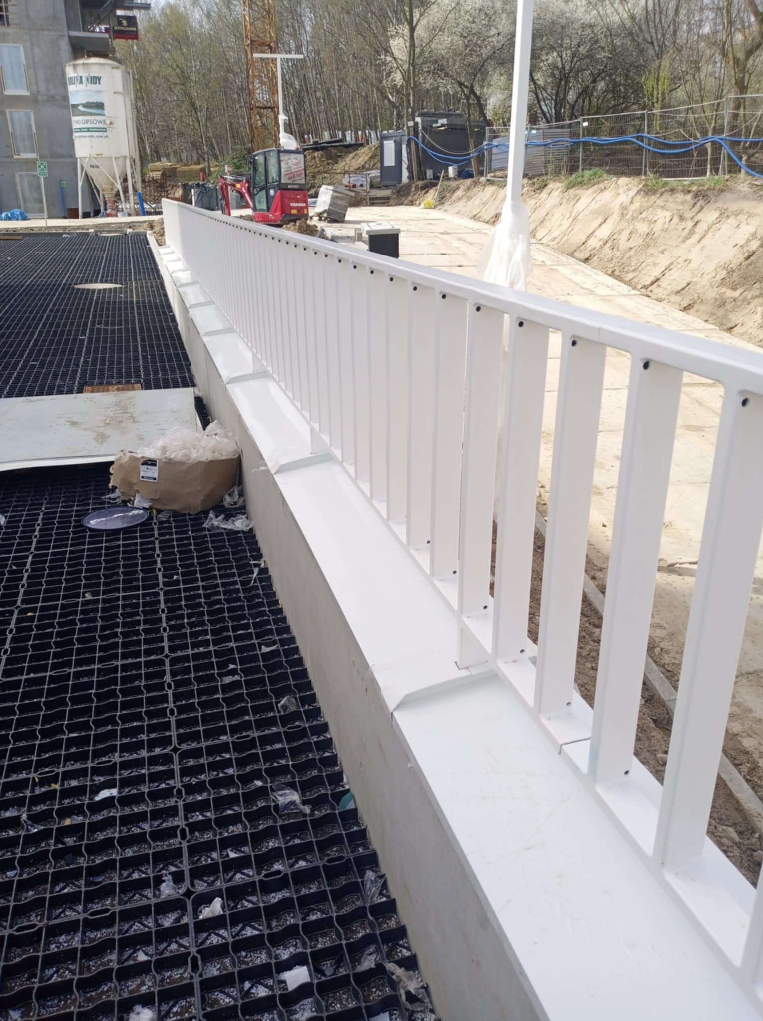 Biała balustrada na betonowym murku, widok z góry na czarną kratę, w tle plac budowy z maszynami i drzewami.