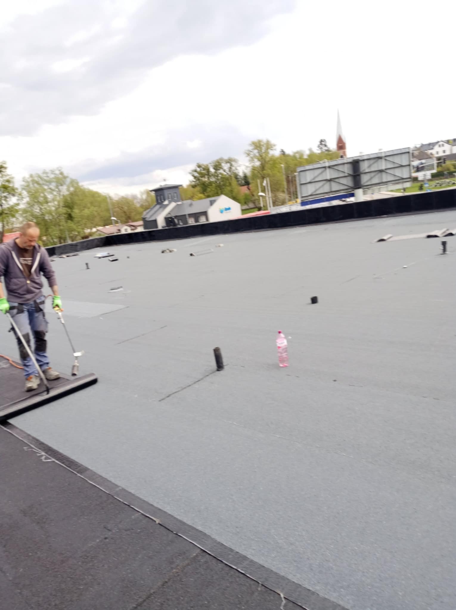 Pracownik w Gdańsku uszczelnia papą płaski dach za pomocą specjalistycznego palnika na kółkach, w tle budynki i billboard.