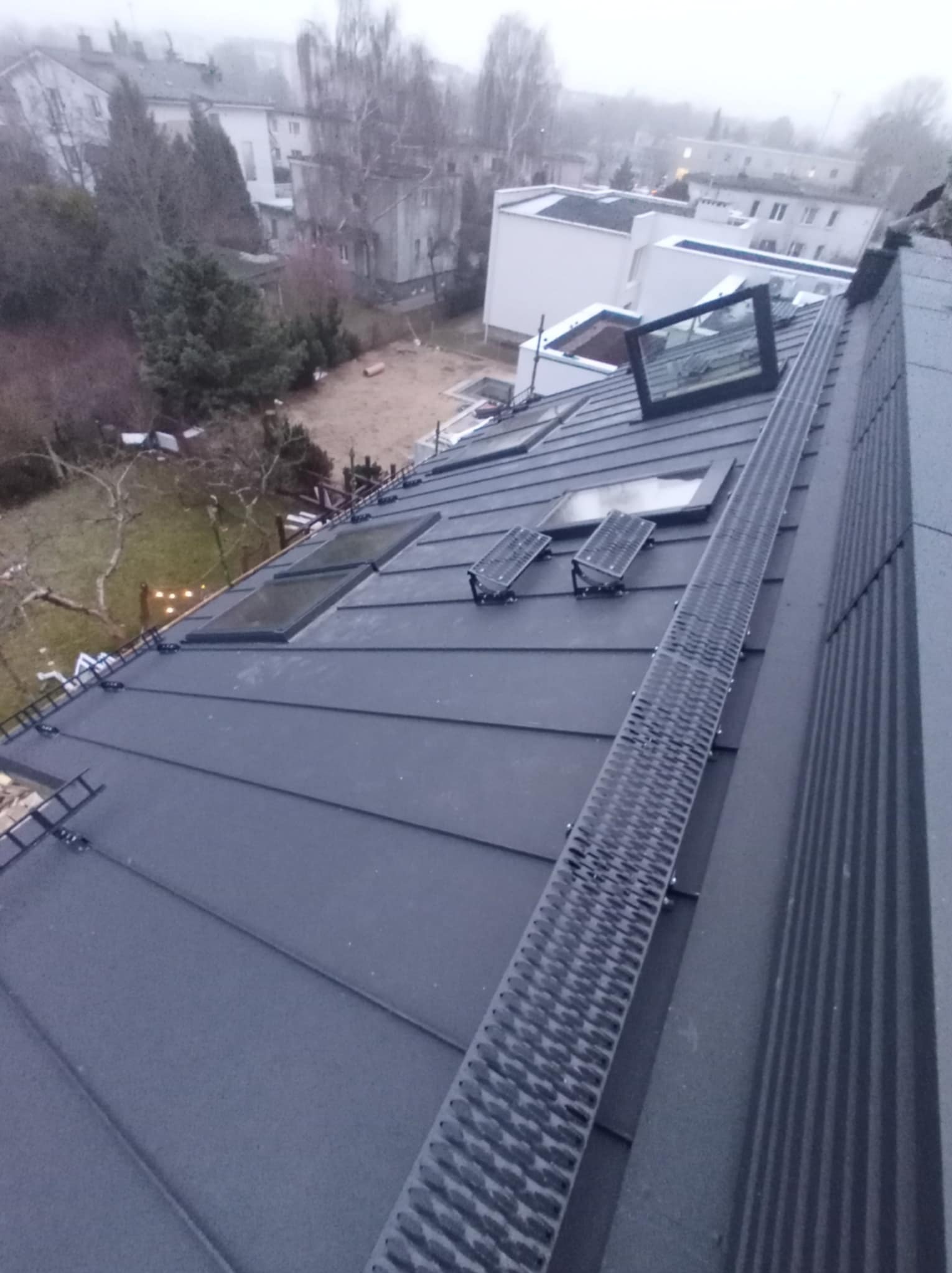 Szary dach z blachy trapezowej z oknami dachowymi i metalową ścieżką komunikacyjną, widok z góry w pochmurny dzień.