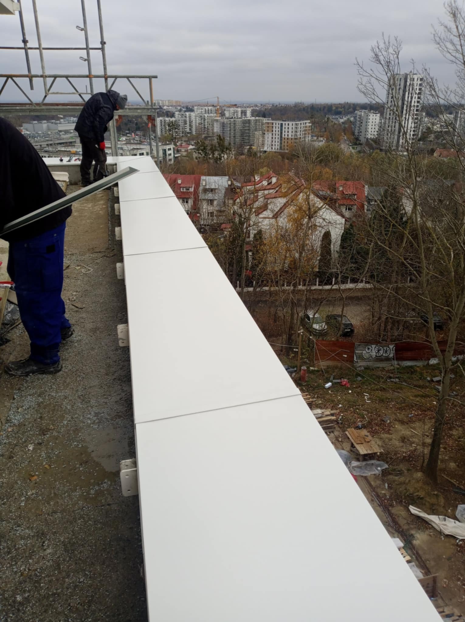 Montaż białych paneli na krawędzi dachu z widokiem na miasto w tle, pracownicy na rusztowaniu, pochmurny dzień.