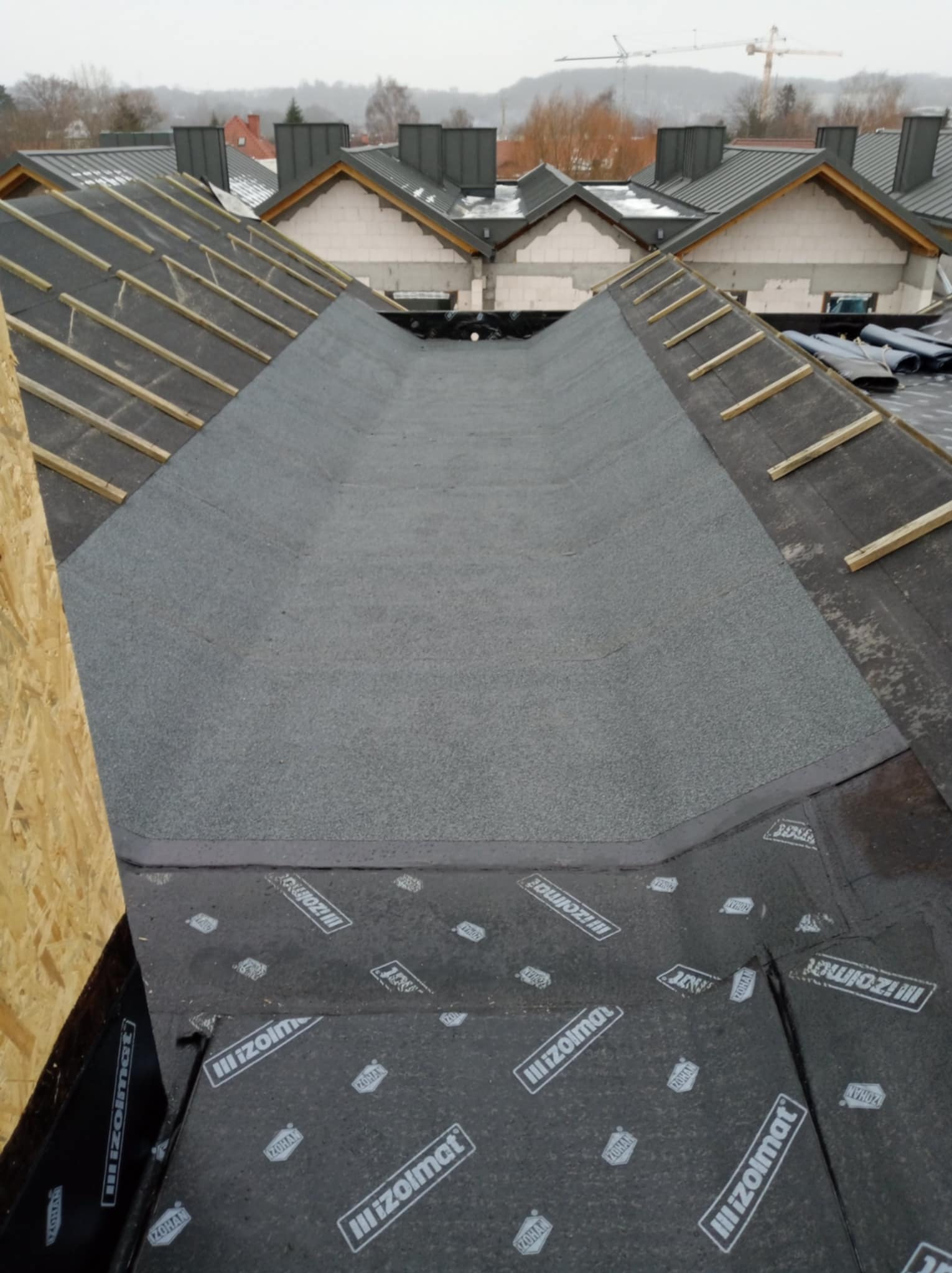 Widok z góry na fragment dachu z papy z logo 'Izolmat', z przygotowaną powierzchnią pod kolejne warstwy, widoczne drewniane łaty i kominy na tle zabudowy miejskiej pod pochmurnym niebem.