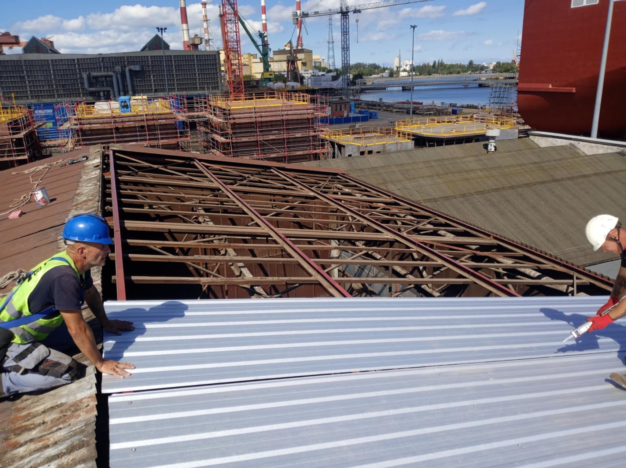Pracownicy w kaskach montują blachę trapezową na drewnianej konstrukcji dachu przemysłowego, w tle widoczne dźwigi portowe i nabrzeże.