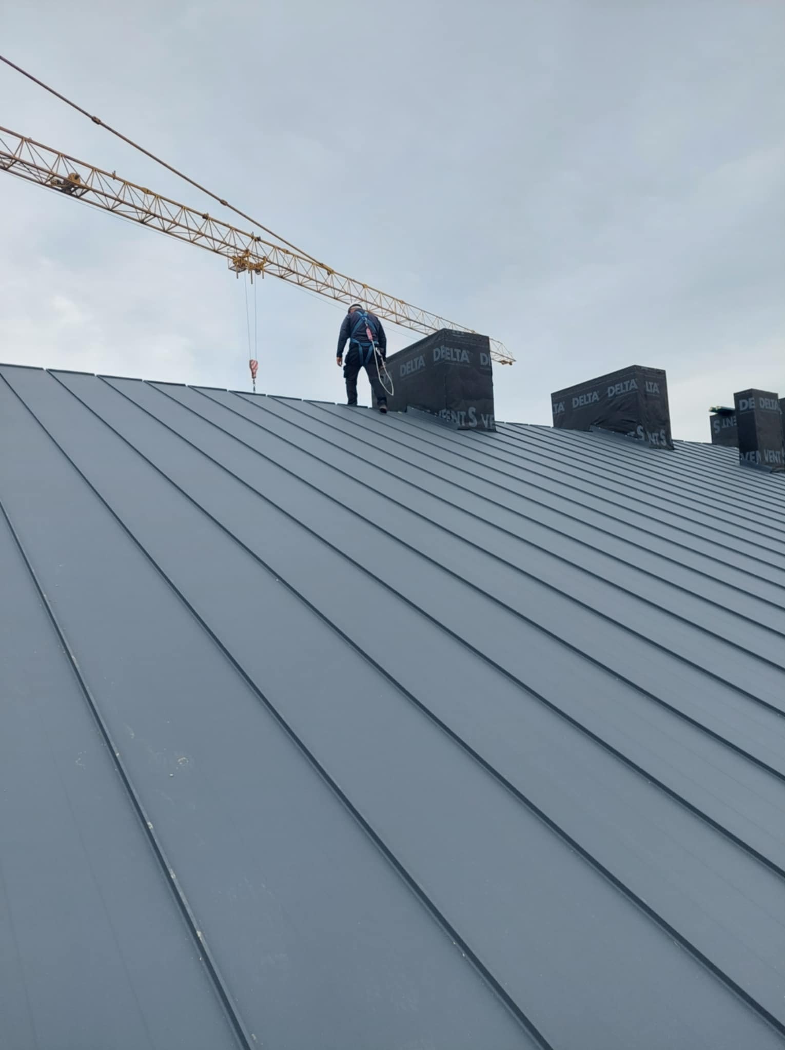 Monter w szelkach bezpieczeństwa stoi na nowym, szarym dachu z blachy na rąbek stojący, w tle widoczny żuraw budowlany i kominki zabezpieczone membraną.