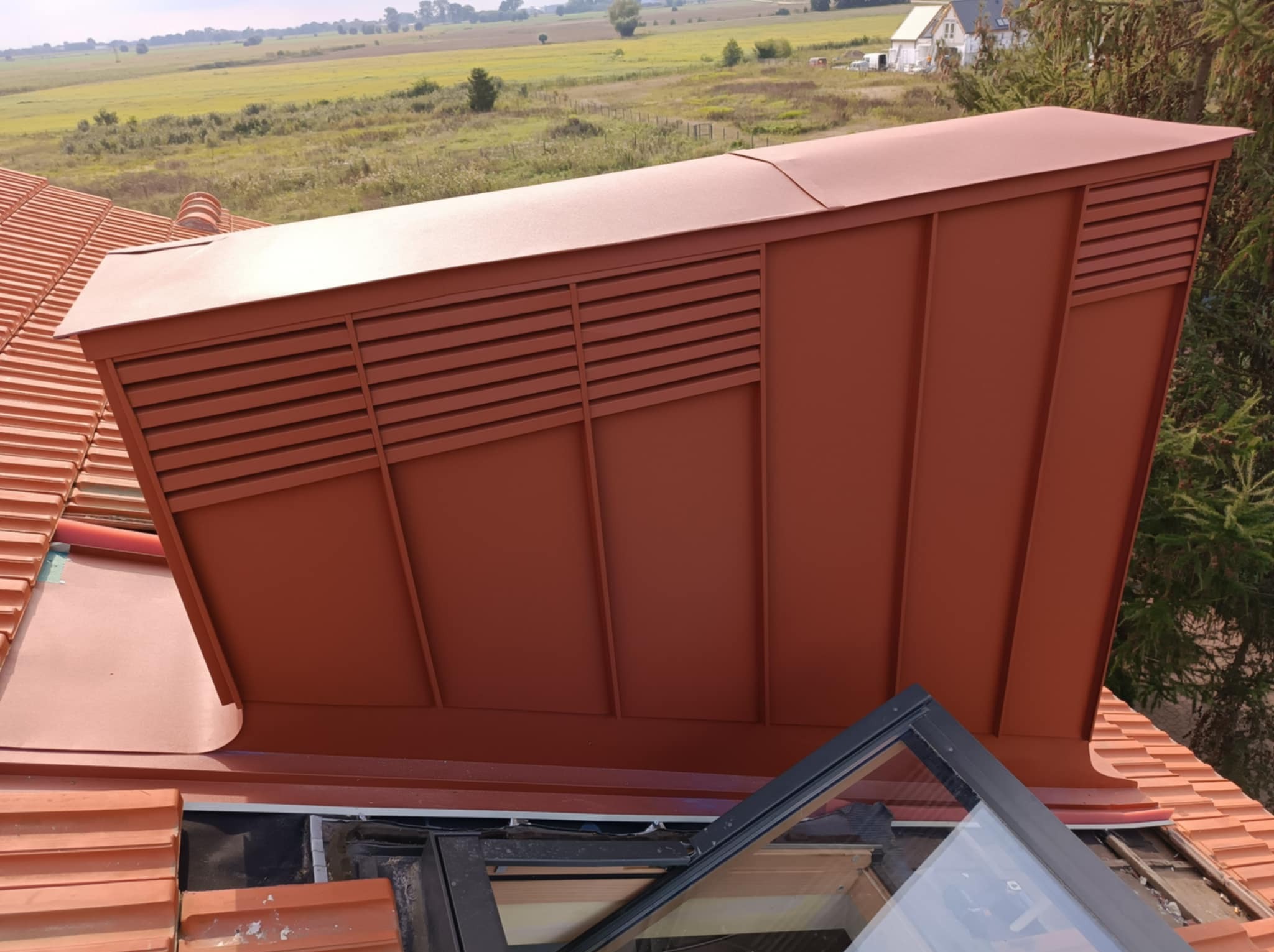 Brązowa obróbka blacharska komina wentylacyjnego na dachu z dachówki ceramicznej, widok z góry, z otwartym oknem dachowym w tle