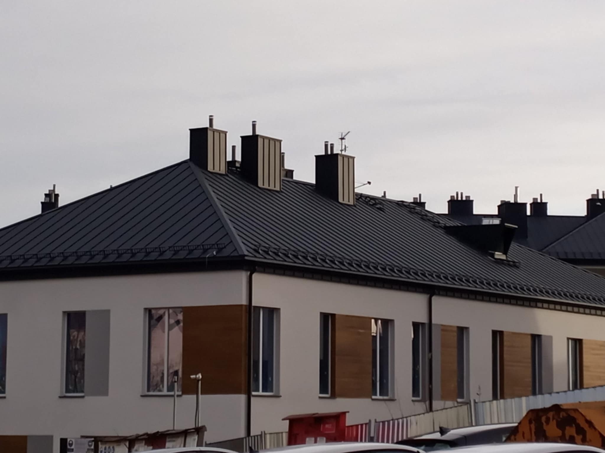 Nowoczesny budynek z ciemnoszarym dachem blaszanym i licznymi kominami, widok z dołu, z fragmentem zaparkowanych samochodów w tle.