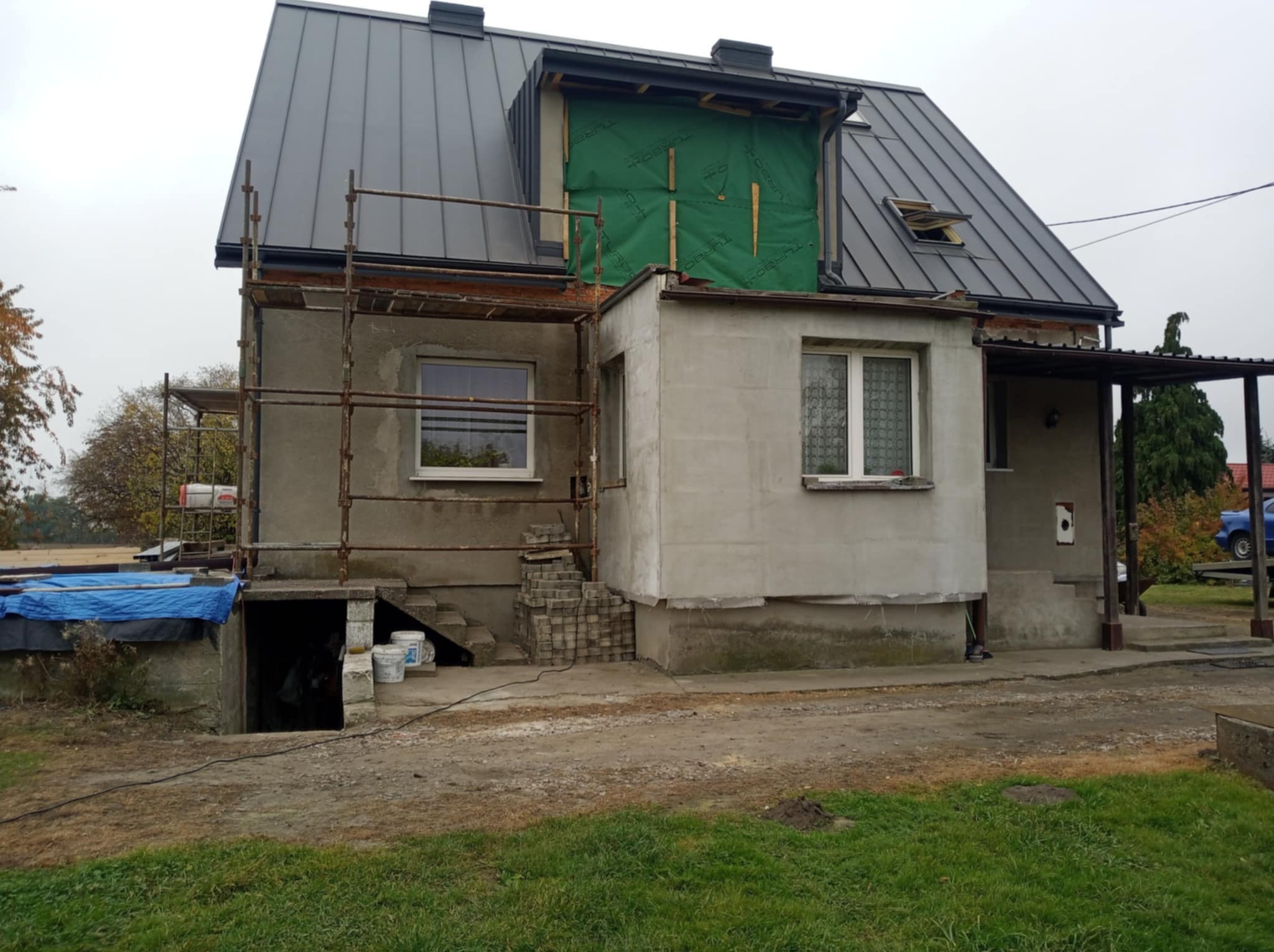 Szary dom jednorodzinny w trakcie remontu dachu, widoczne rusztowania, okno dachowe i fragment membrany dachowej na lukarnie.