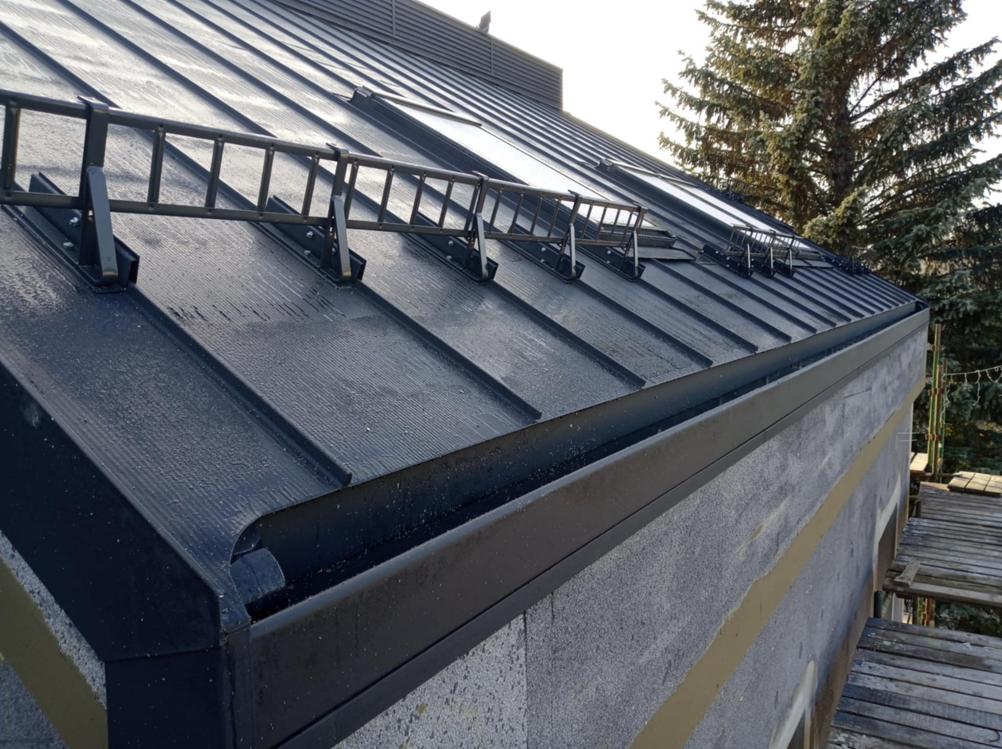 Czarny, metalowy dach z zamontowanymi zabezpieczeniami przeciwśniegowymi w formie drabinki, widoczna również rynna i fragment ocieplonej ściany budynku.