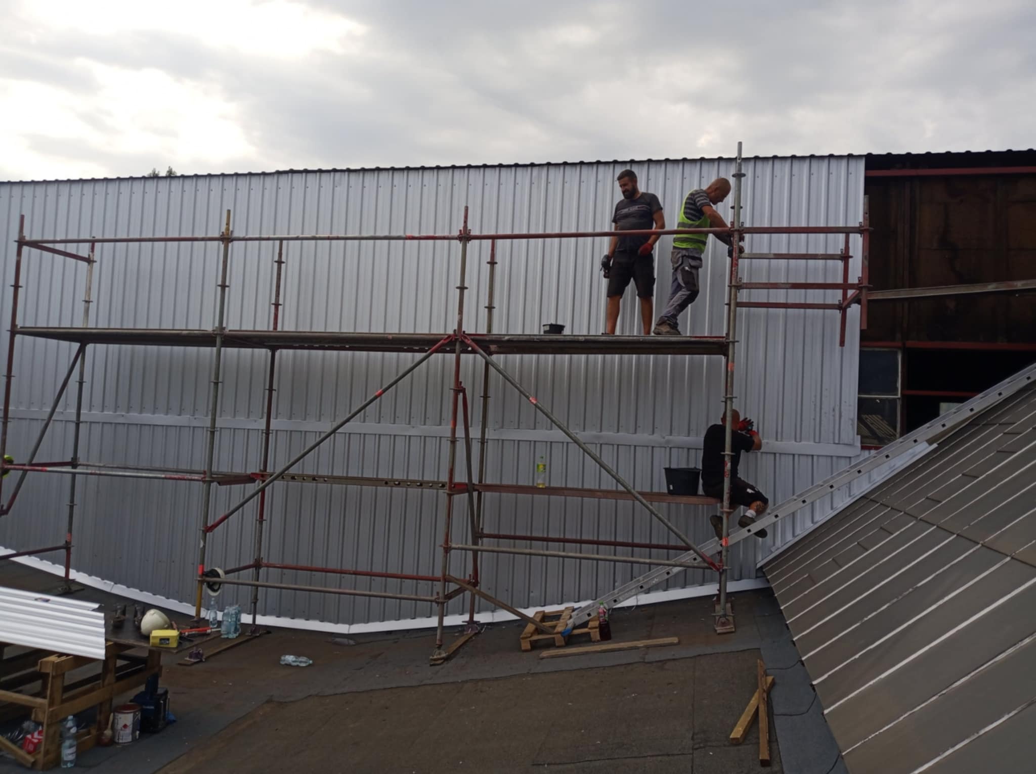 Pracownicy na rusztowaniu montują metalowe panele ścienne, widoczna drabina oparta o dach, narzędzia i materiały budowlane na dachu, pochmurne niebo w tle.