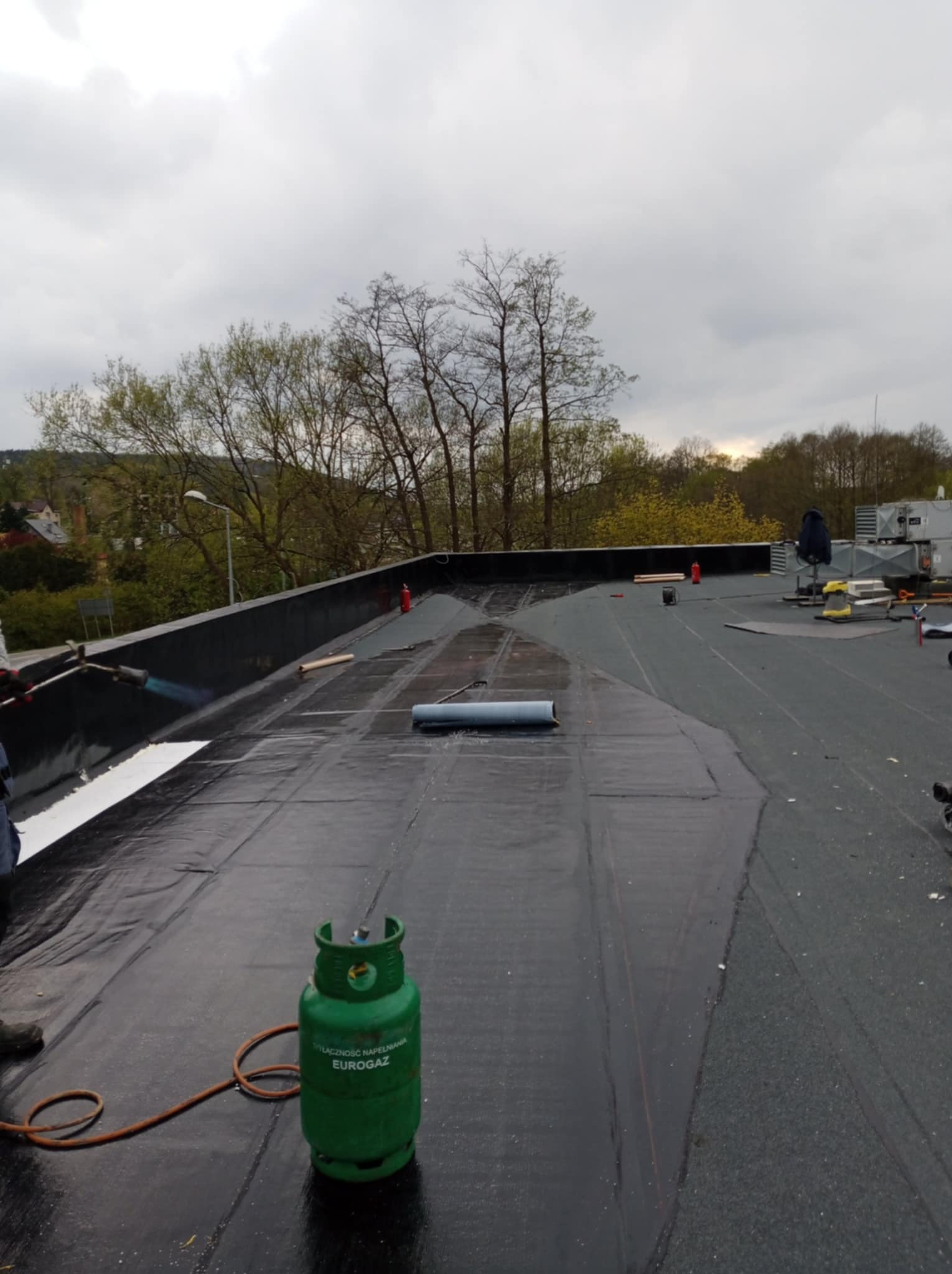 Prace na płaskim dachu pokrytym czarną papą termozgrzewalną; widoczna butla gazowa Eurogaz, palnik dekarski w użyciu, częściowo położona biała warstwa izolacyjna, oraz narzędzia i materiały...