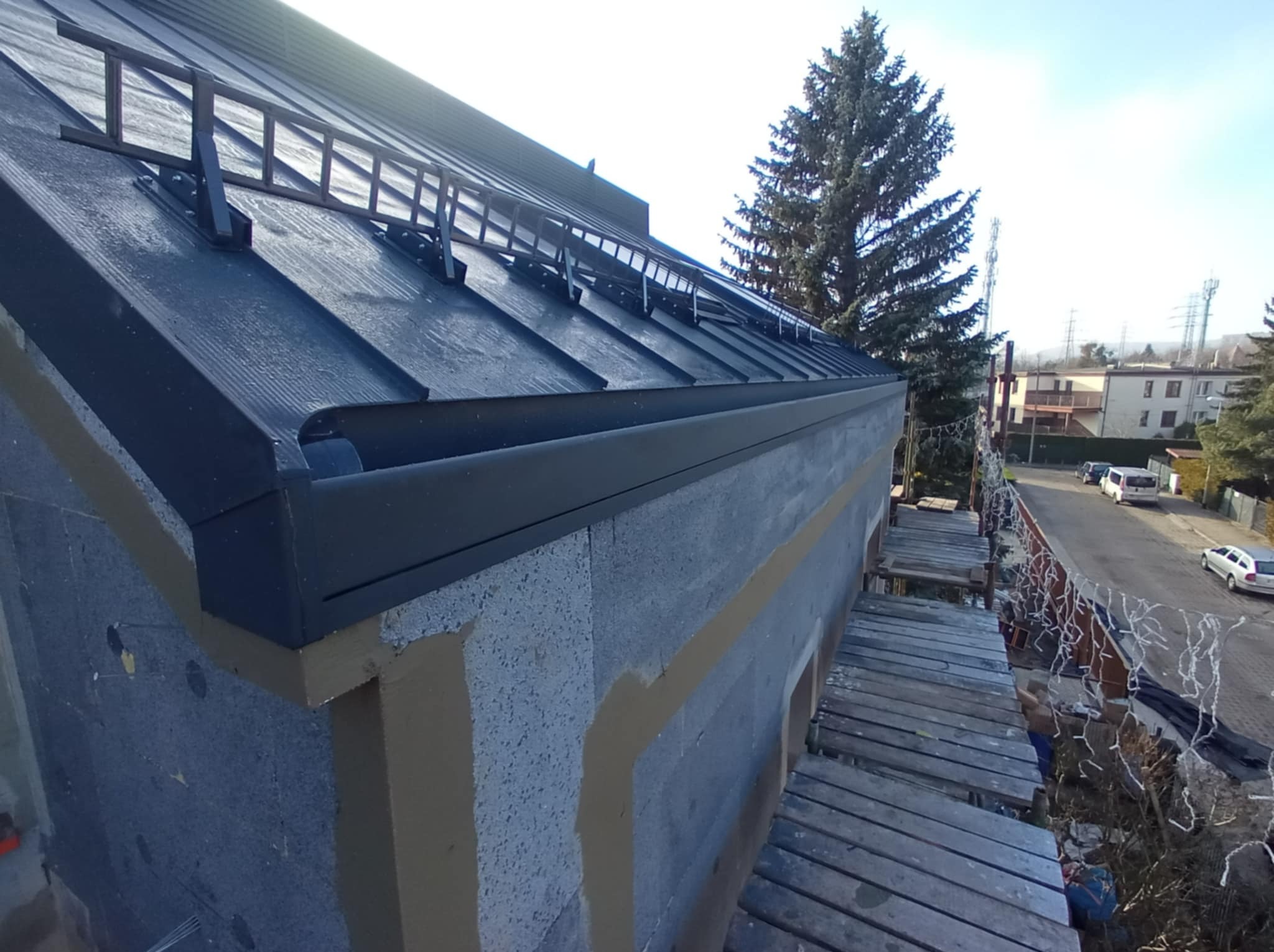 Montaż czarnej blachy na dachu z zamontowanymi drabinkami przeciwśniegowymi, widoczne rusztowanie i ocieplenie budynku w trakcie prac wykończeniowych.