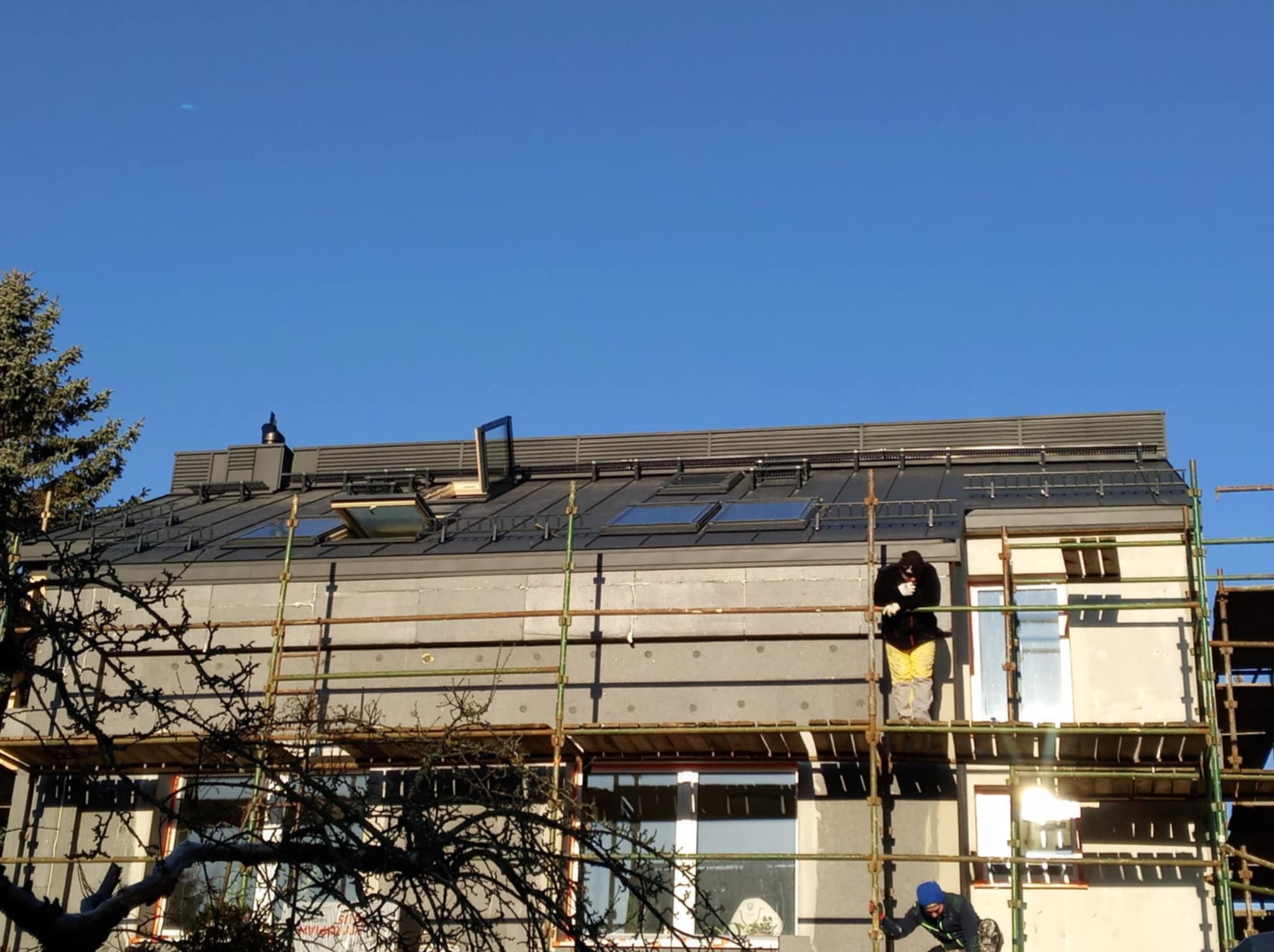 Dwóch robotników na rusztowaniu przy ocieplonym budynku z szarym dachem z kilkoma oknami dachowymi, widok z dołu na tle błękitnego nieba.