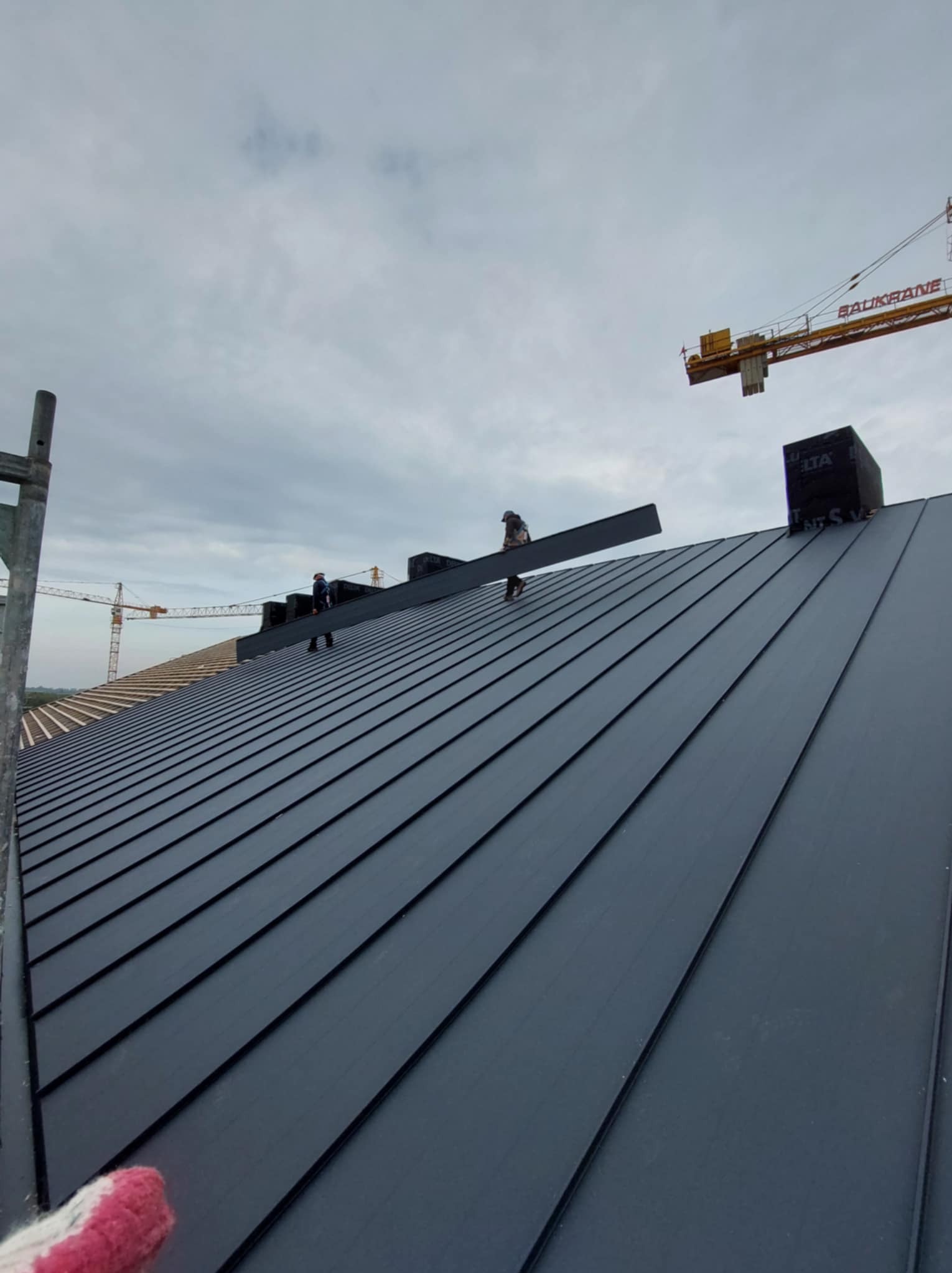 Montaż blachy na dachu z widocznymi pracownikami, dźwigiem budowlanym w tle i częściowo odkrytą konstrukcją dachu