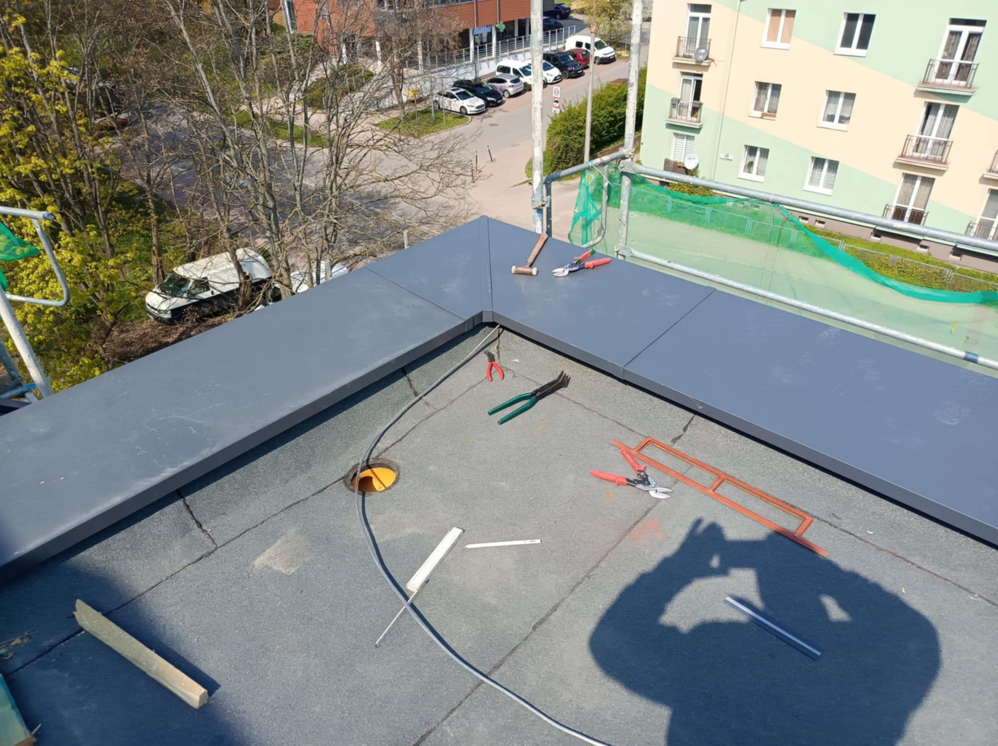 Płaski dach pokryty papą z widocznym fragmentem obróbki blacharskiej, narzędziami dekarskimi (młotek, nożyce do blachy) i otworem wentylacyjnym w tle zabudowa miejska.