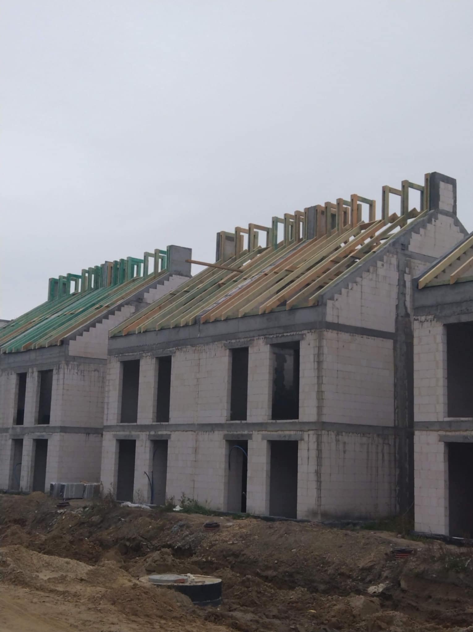 Surowa konstrukcja szeregówki z widoczną więźbą dachową w trakcie budowy, z elementami zaimpregnowanego drewna na niektórych dachach, na tle pochmurnego nieba.