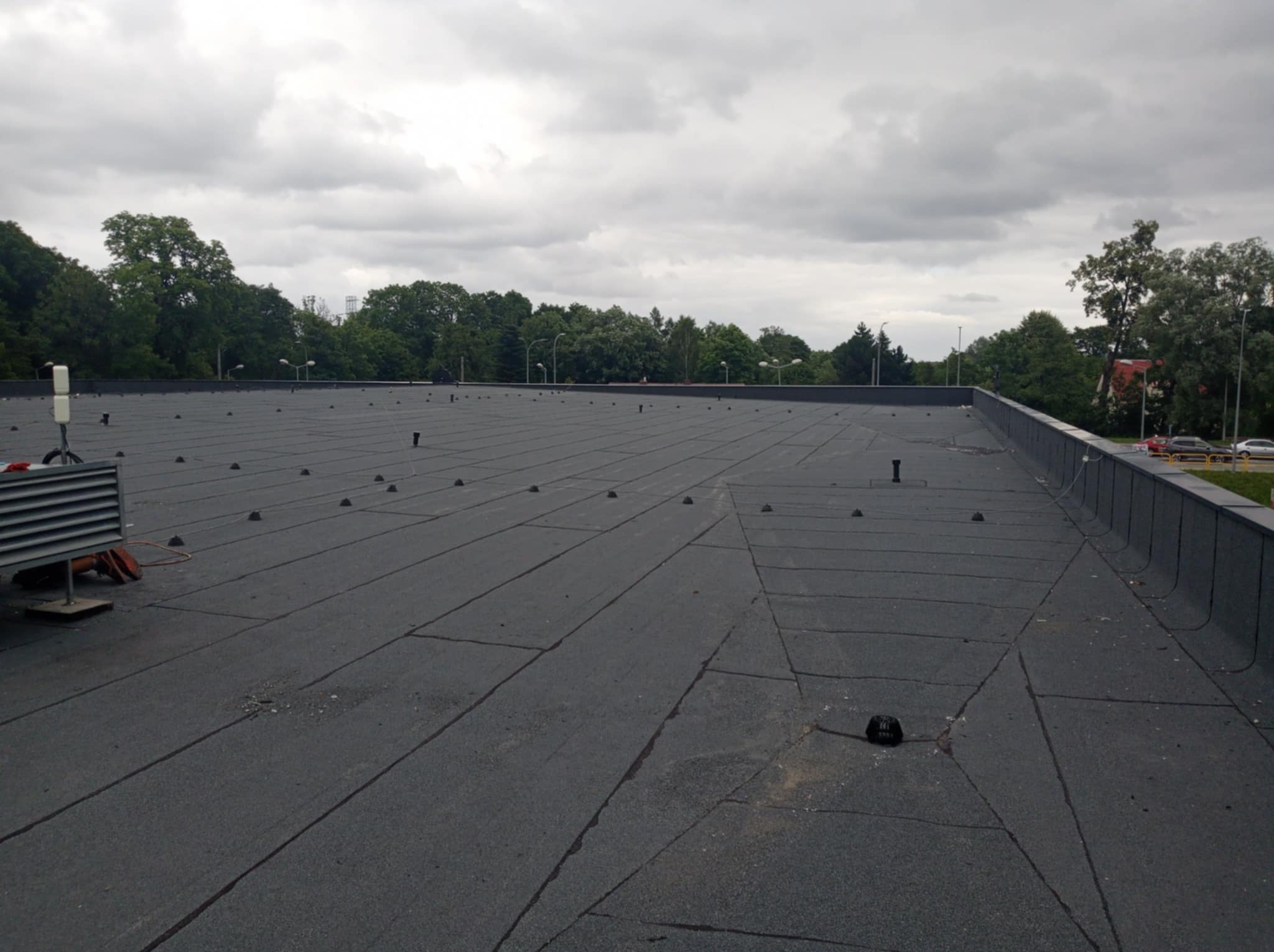 Płaski dach pokryty papą termozgrzewalną z widocznymi kominkami wentylacyjnymi i krawędzią dachu, w tle zieleń drzew i pochmurne niebo.