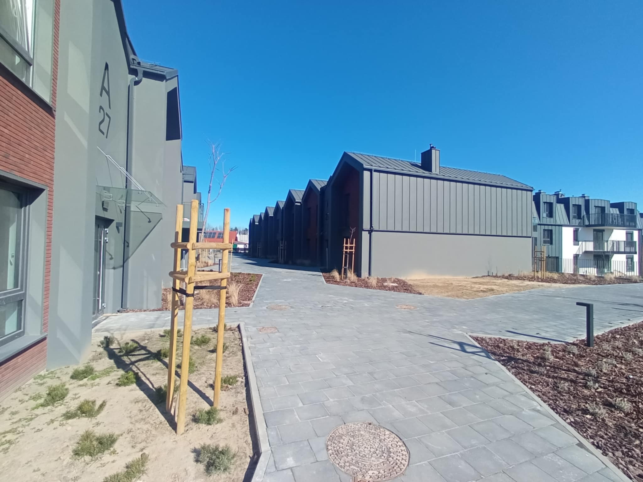 Szereg identycznych, nowoczesnych budynków z szarymi, metalowymi dachami w słoneczny dzień, widok z poziomu chodnika z kostki brukowej.