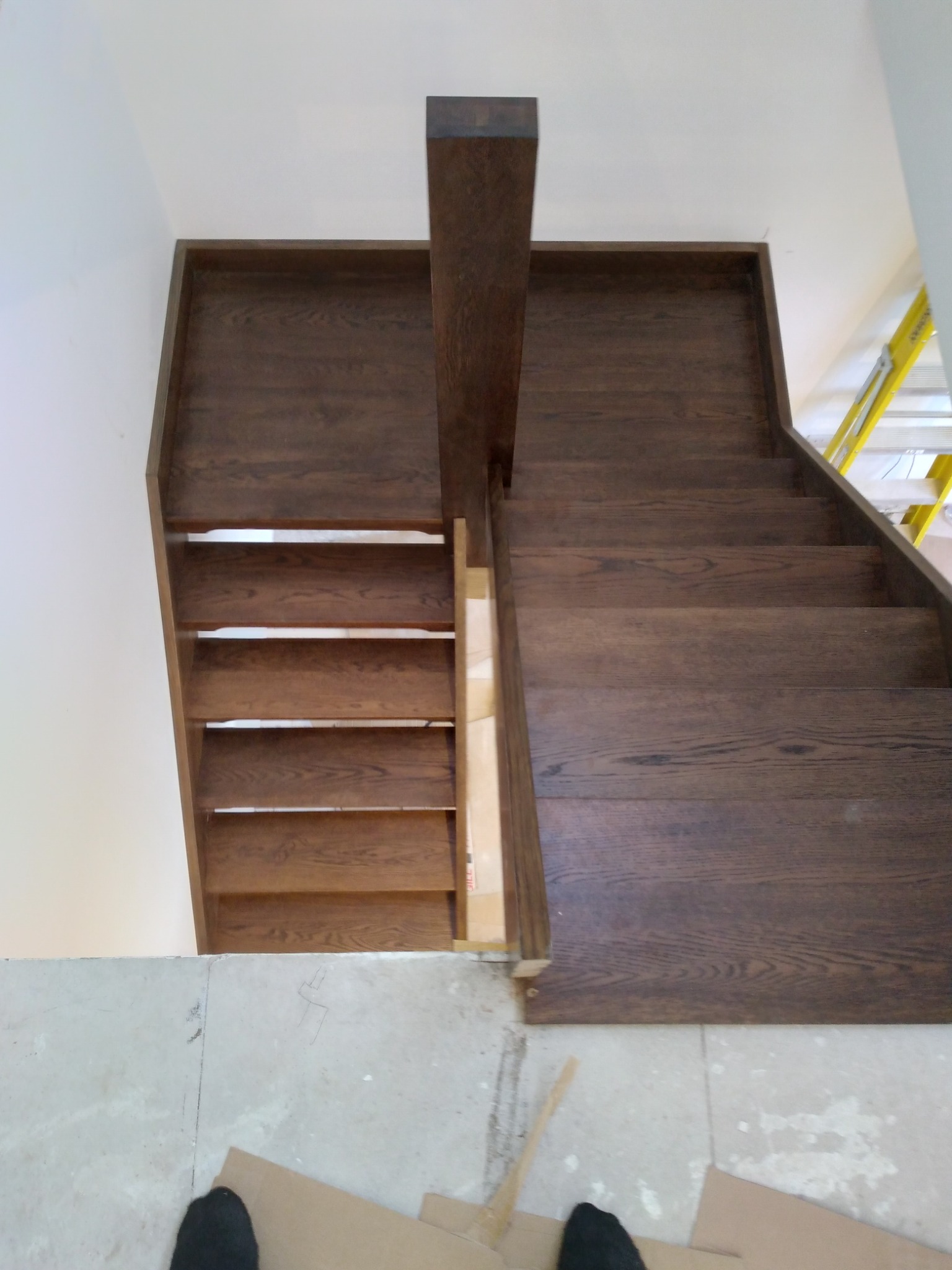 Montaż drewnianych schodów z ciemnego drewna, widok z góry na nieukończoną konstrukcję, z widocznymi elementami montażowymi i fragmentem drabiny w tle.