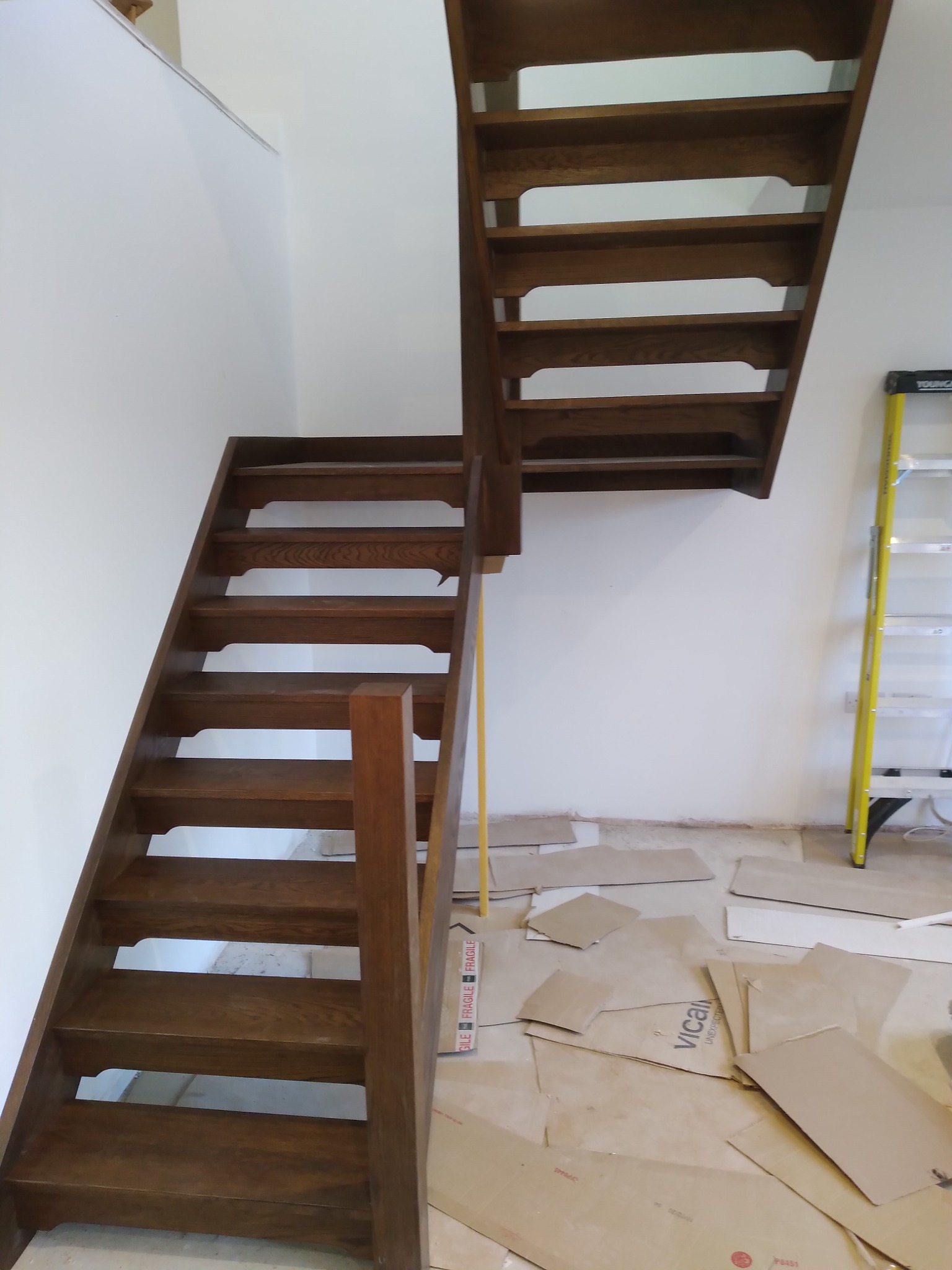 Drewniane schody z balustradą, widok z dołu, na podłodze rozłożone kartony zabezpieczające podczas montażu, w tle drabina aluminiowa.