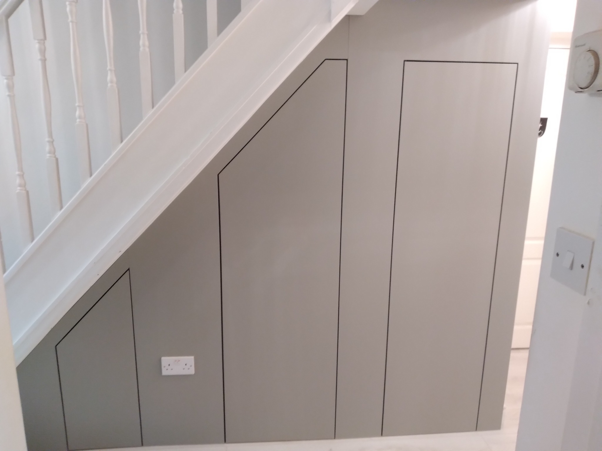 Szare, minimalistyczne drzwi do schowka pod schodami z czarnymi obrysami, widoczne białe schody oraz gniazdko elektryczne.