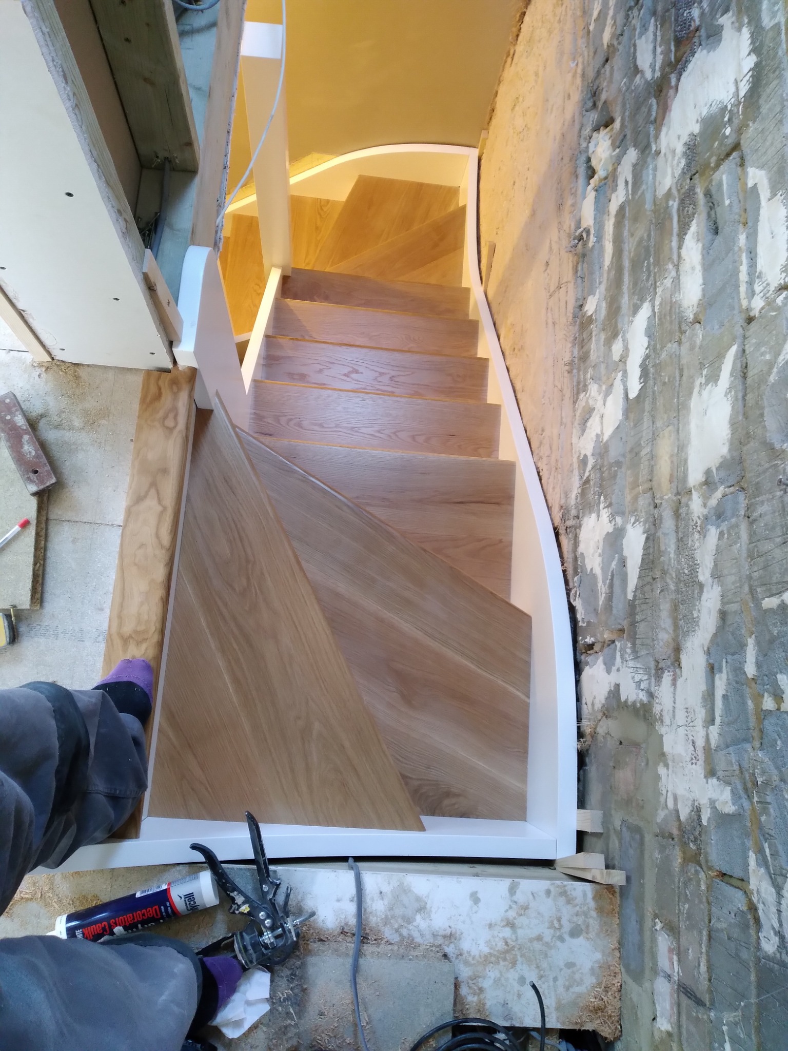 Montaż drewnianych schodów z białymi balustradami w surowym stanie wykończenia, widoczne narzędzia i fragment osoby w skarpetkach.