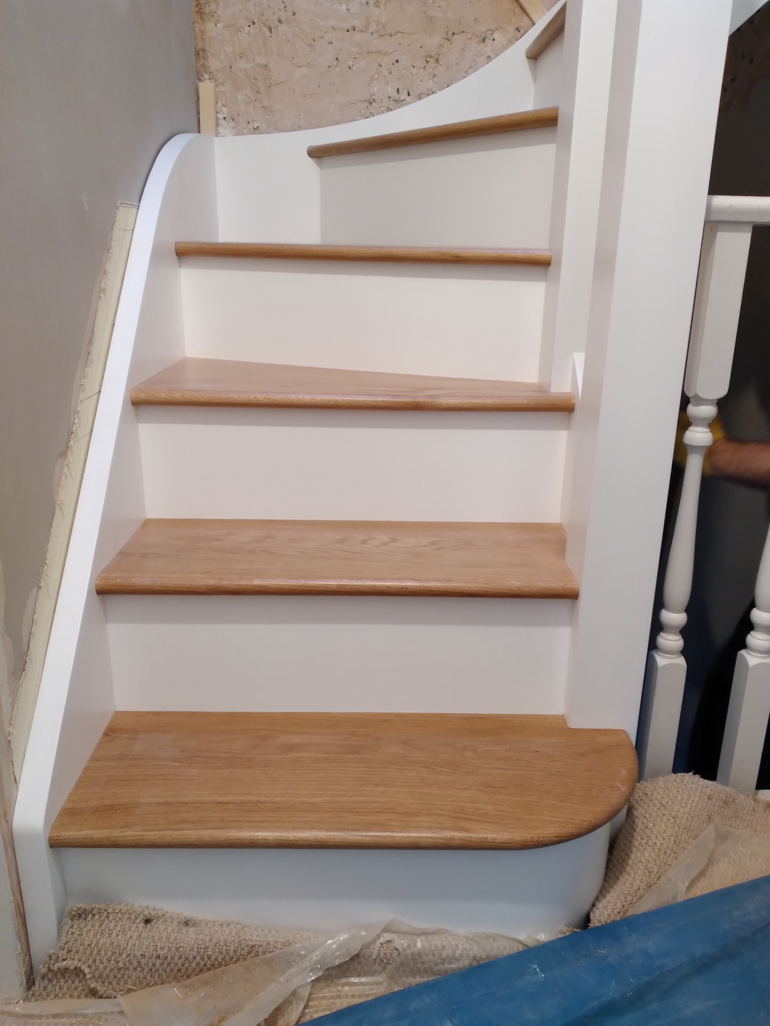 Schody drewniane z białymi podstopnicami i balustradą, widok z dołu, w trakcie montażu, z widocznymi niedoskonałościami na ścianie.