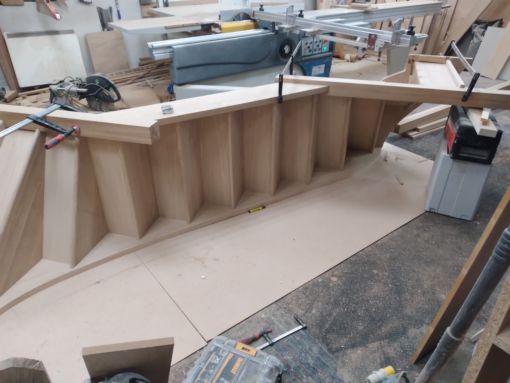 Wnętrze warsztatu stolarskiego: etap budowy drewnianej konstrukcji, prawdopodobnie szafy wnękowej, z widocznymi narzędziami i maszynami stolarskimi w tle.