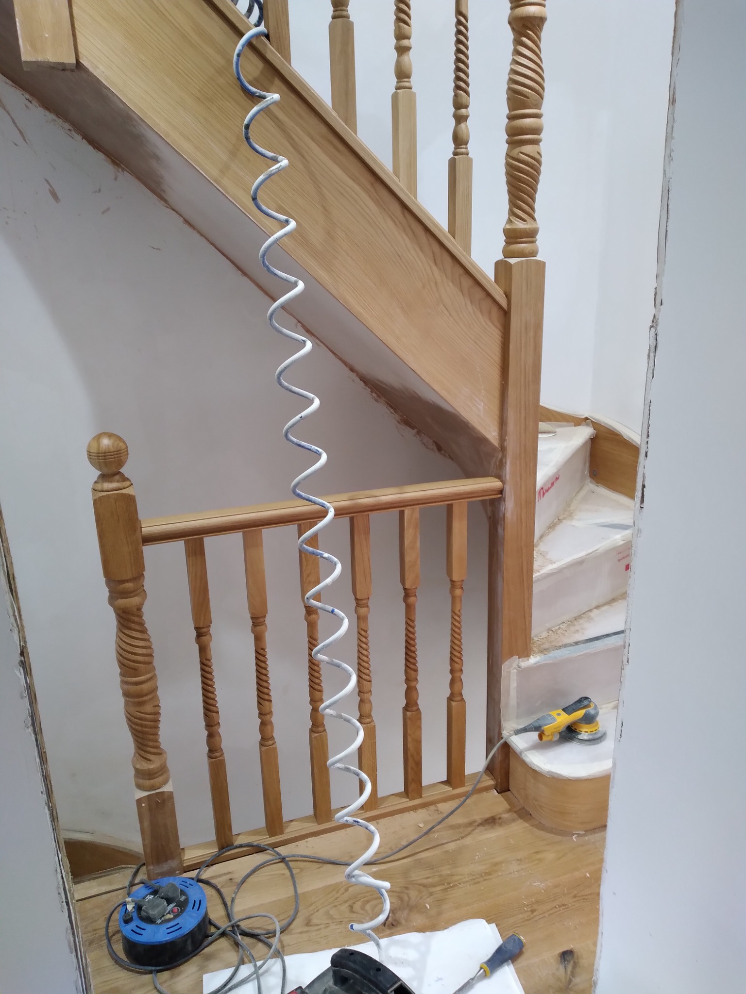 Drewniane schody z toczonymi balustradami w trakcie renowacji, z widoczną szlifierką oscylacyjną i spiralnym przewodem pneumatycznym, na podłodze leżą narzędzia i przedłużacz.