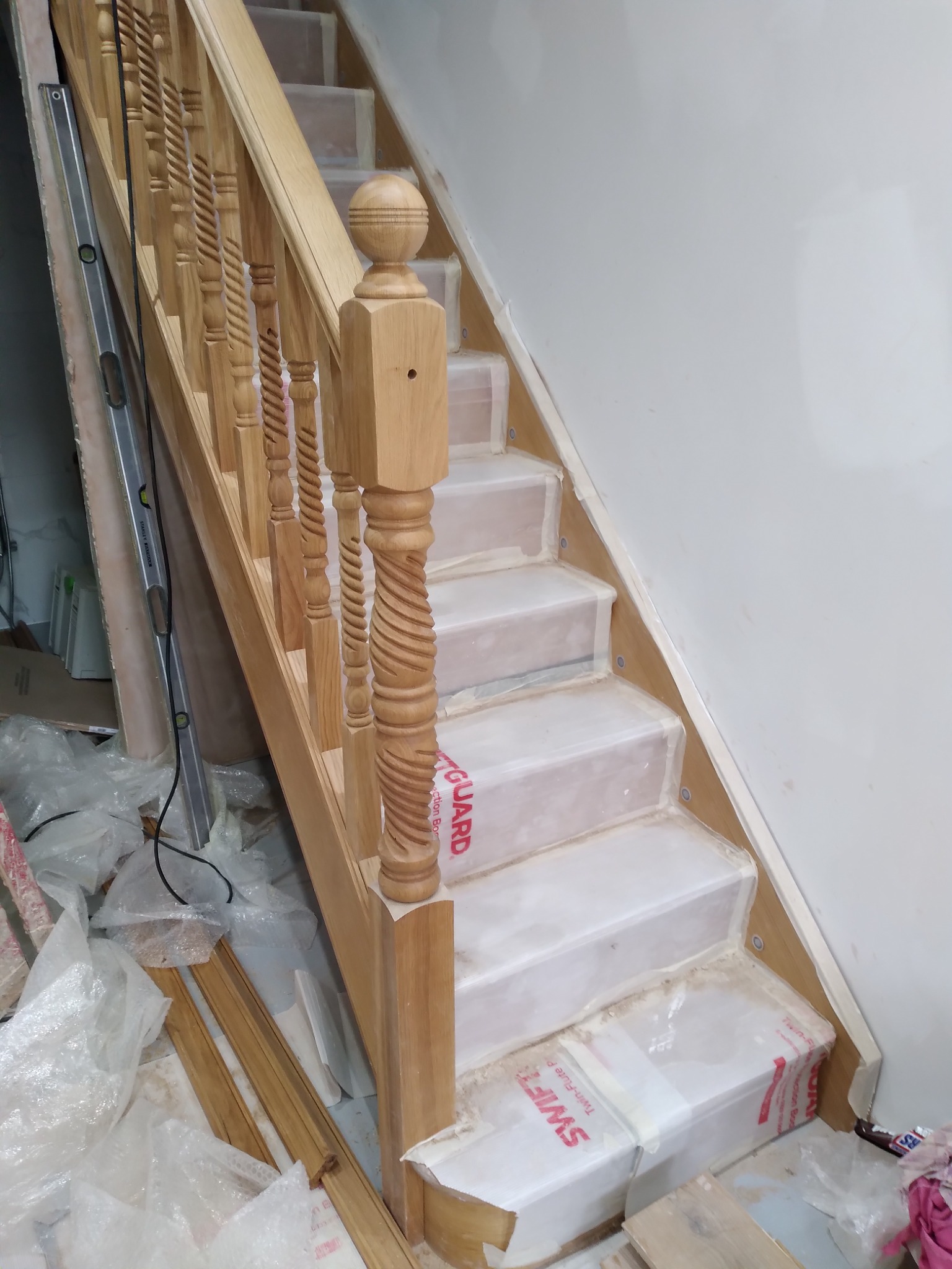 Drewniane schody z balustradą o toczonych tralkach w trakcie montażu, zabezpieczone folią, widoczna poziomica i resztki materiałów budowlanych.