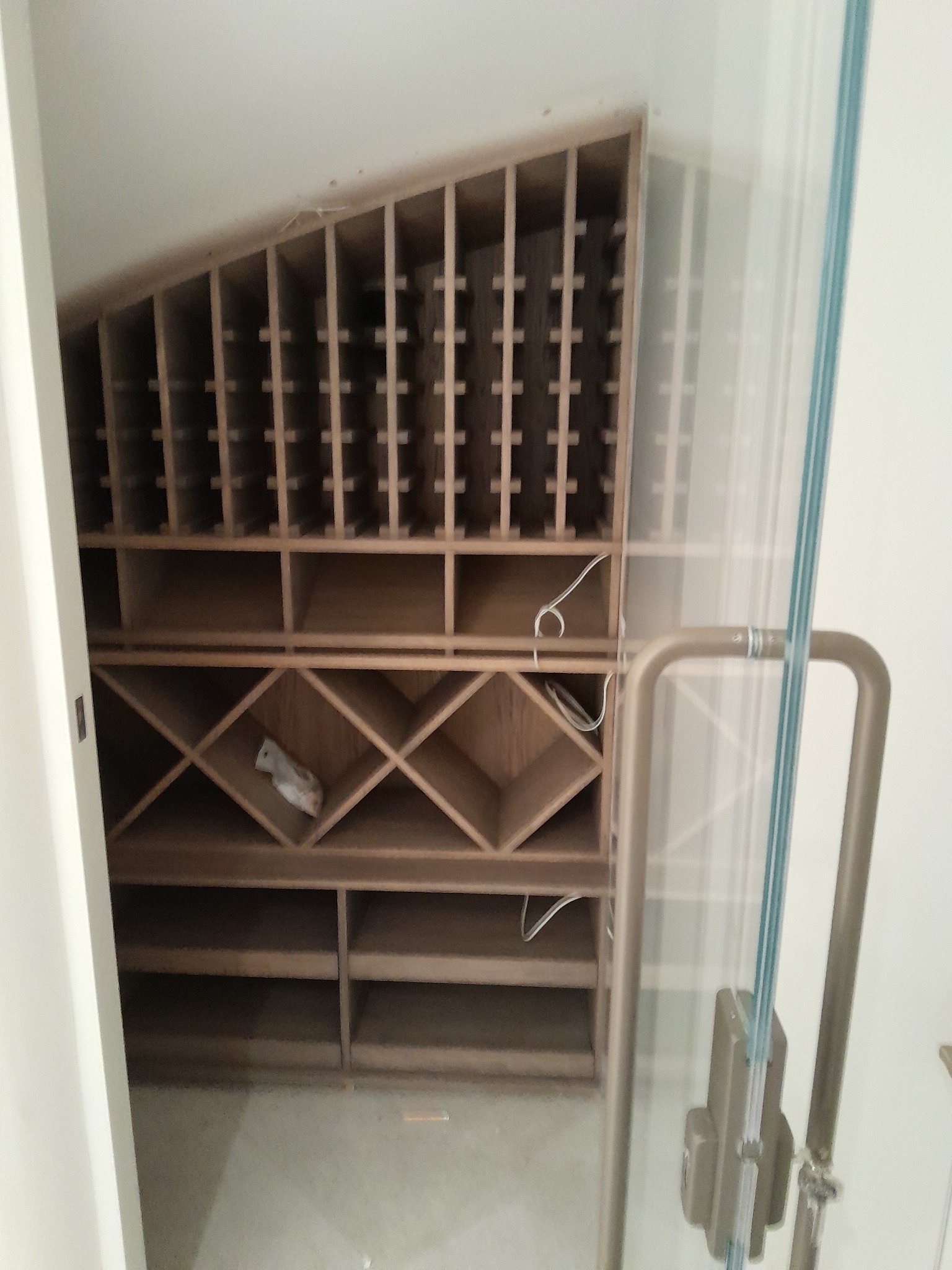 Drewniany regał z półkami o różnej wielkości, w tym ukośne półki na butelki, widoczny przez szklane drzwi z metalowym uchwytem.