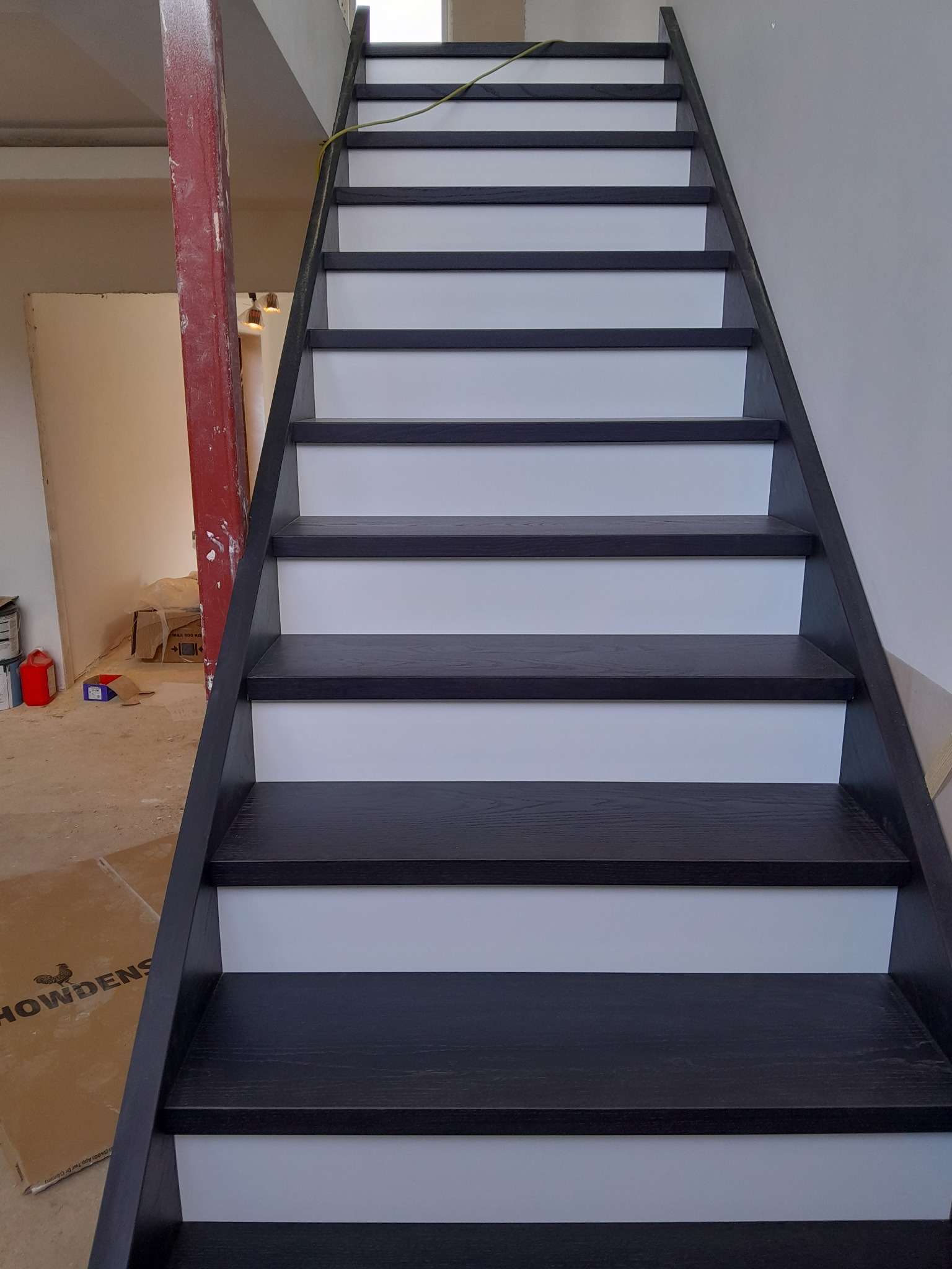 Schody drewniane z czarnymi stopniami i białymi podstopnicami, widok z dołu w trakcie montażu, z widocznym kartonem na podłodze i elementami konstrukcyjnymi w tle.
