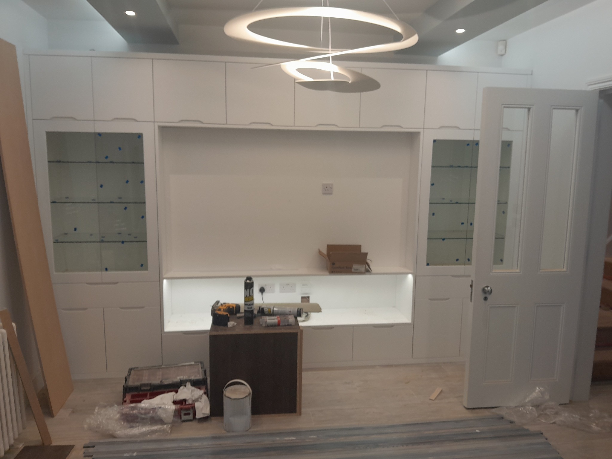 Biała zabudowa meblowa z przeszklonymi witrynami i otwartymi półkami w trakcie montażu, widoczne narzędzia i materiały budowlane, nowoczesna lampa sufitowa.
