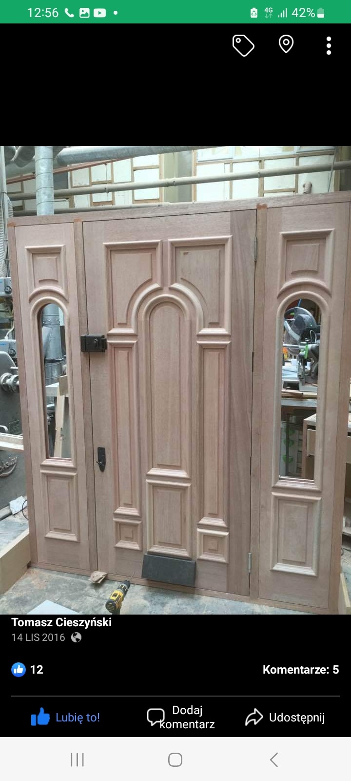 Surowe drewniane drzwi wejściowe z dekoracyjnymi panelami, w trakcie montażu, widoczna wiertarka i tło warsztatowe.