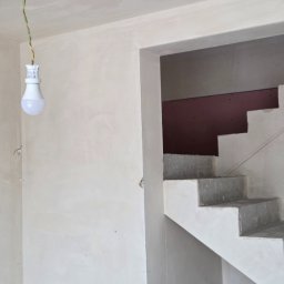PRO100TYNKI - Ściana po nałożeniu gładzi gipsowej, widoczne schody w stanie surowym i żarówka na kablu. Prace wykończeniowe w nowym budynku.