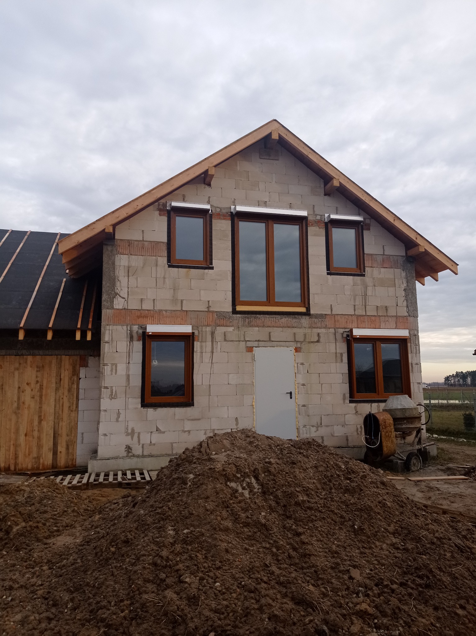 Surowy stan budynku z pustaków, z zamontowanymi oknami z brązowymi ramami i roletami, widoczna konstrukcja dachu, obok sterta ziemi i betoniarka.